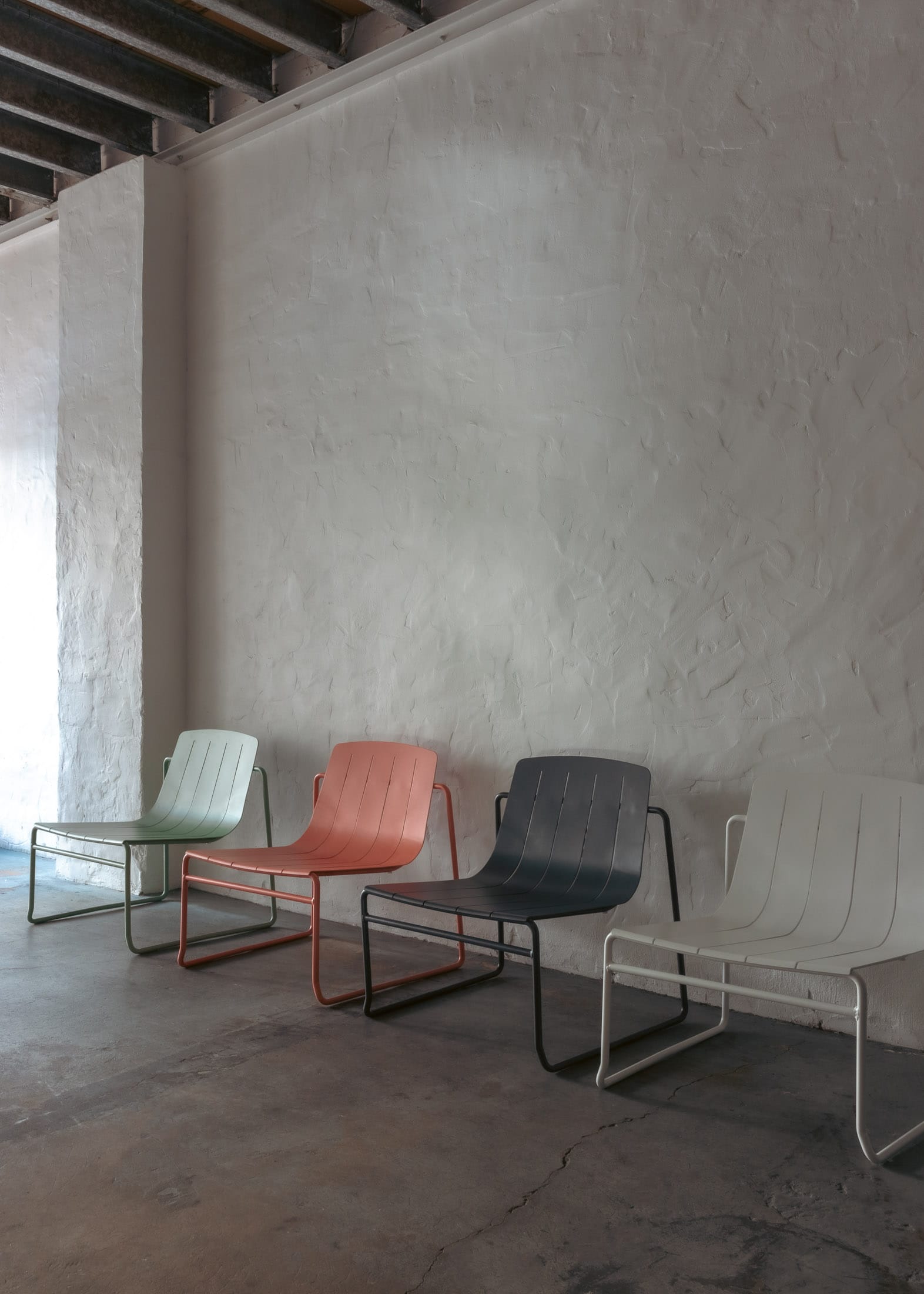Franka's Frankie Outdoor Lounge Chairs.Four unique outdoor lounge chairs presented in a row against a textured white wall. Each chair features a different color - mint green, coral, grey, and white - and rests on slender, rounded metal frames.