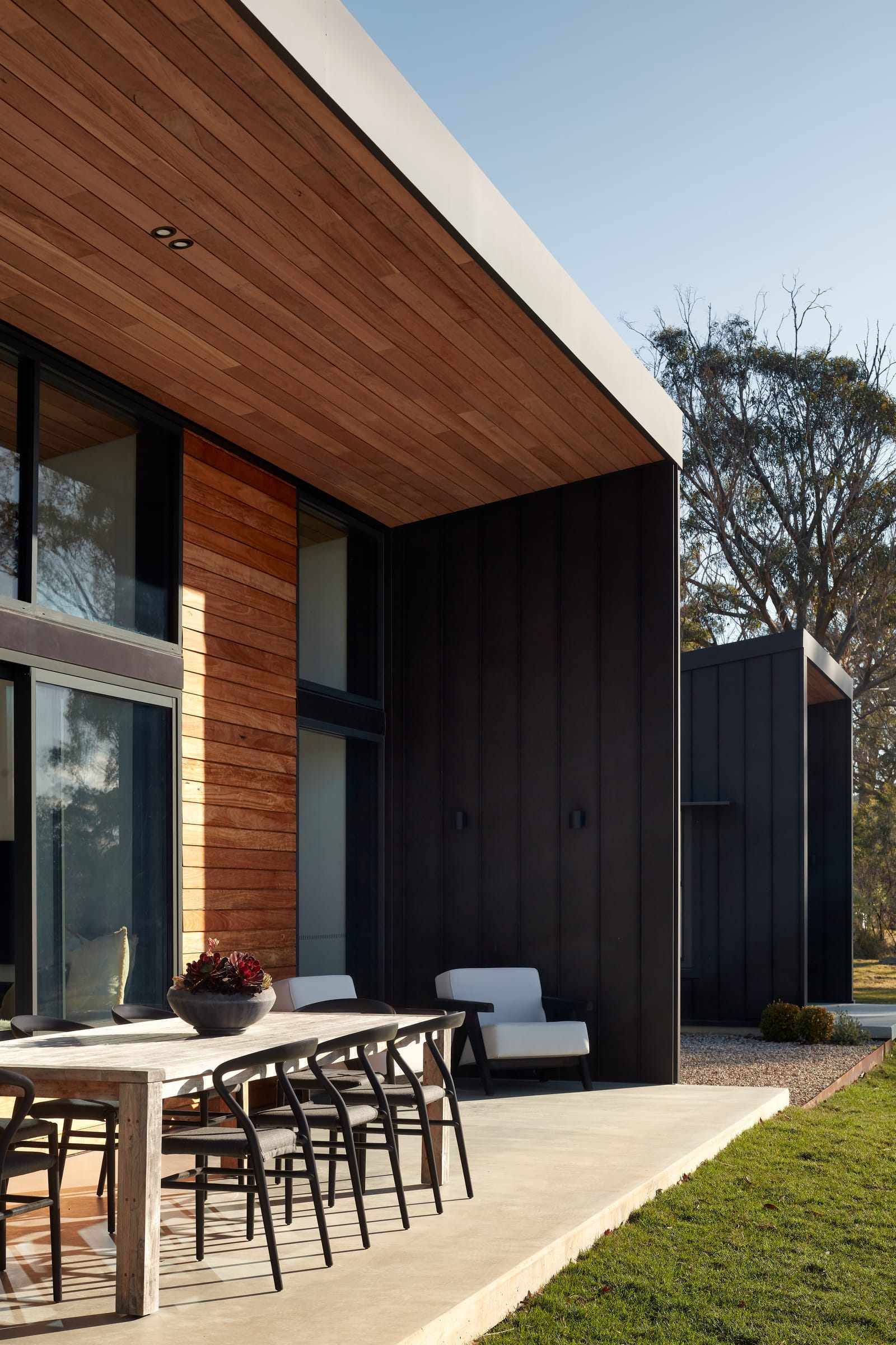 An exterior view of House Woodland by AD Design Studio, highlighting the modern architectural design with a mix of materials. The facade combines wood siding with black metal panels. The house has large windows and sliding doors, integrating indoor and outdoor spaces. A wooden deck extends from the house, furnished with an outdoor dining area.