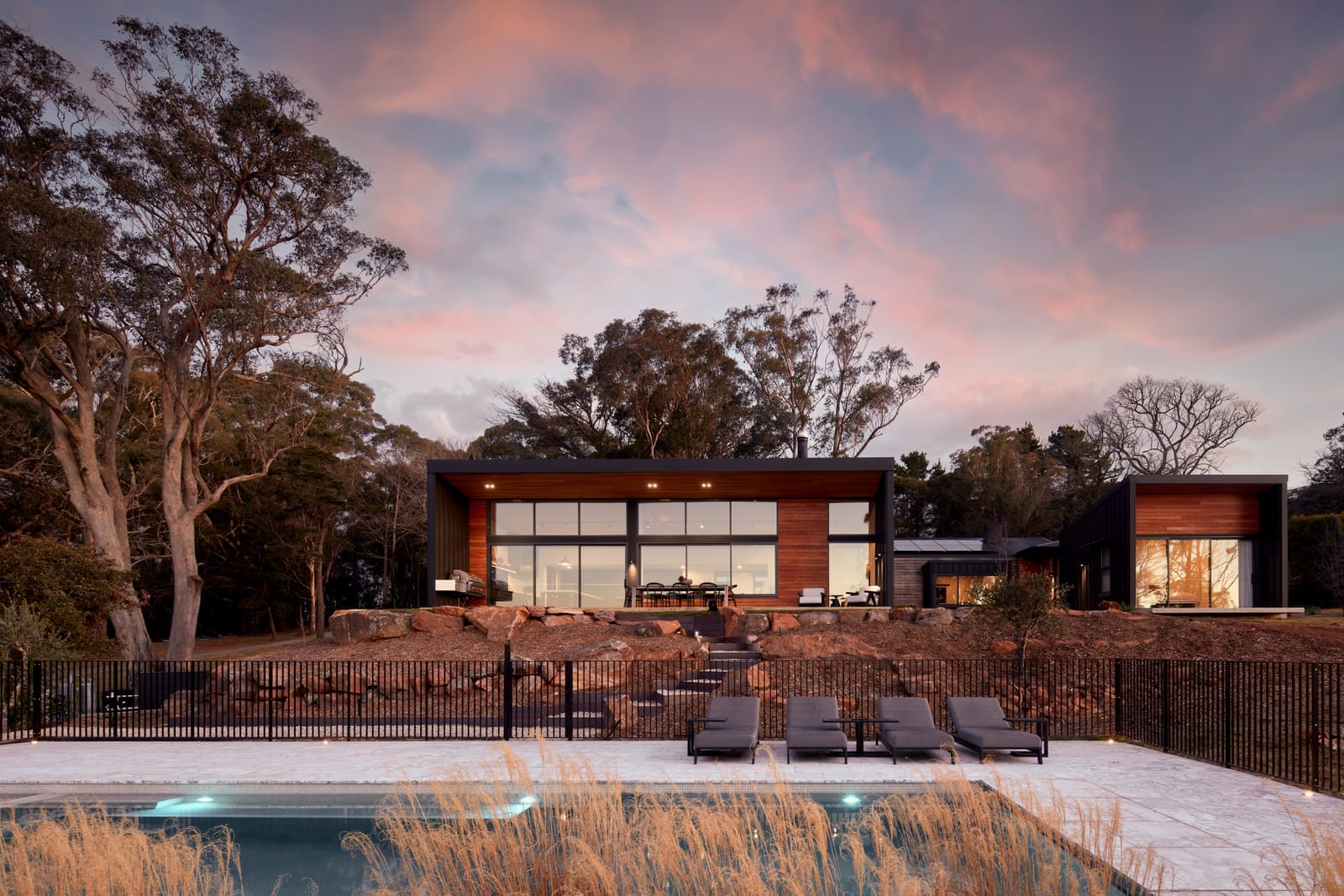 House Woodland by AO Design Studio. he image presents a twilight view of House Woodland by AD Design Studio, with the interior lights casting a warm glow against the dusky sky. The modern house has a flat roof and large windows that showcase the interior. The outdoor area includes a patio with a dining set and a pool that reflects the evening sky.