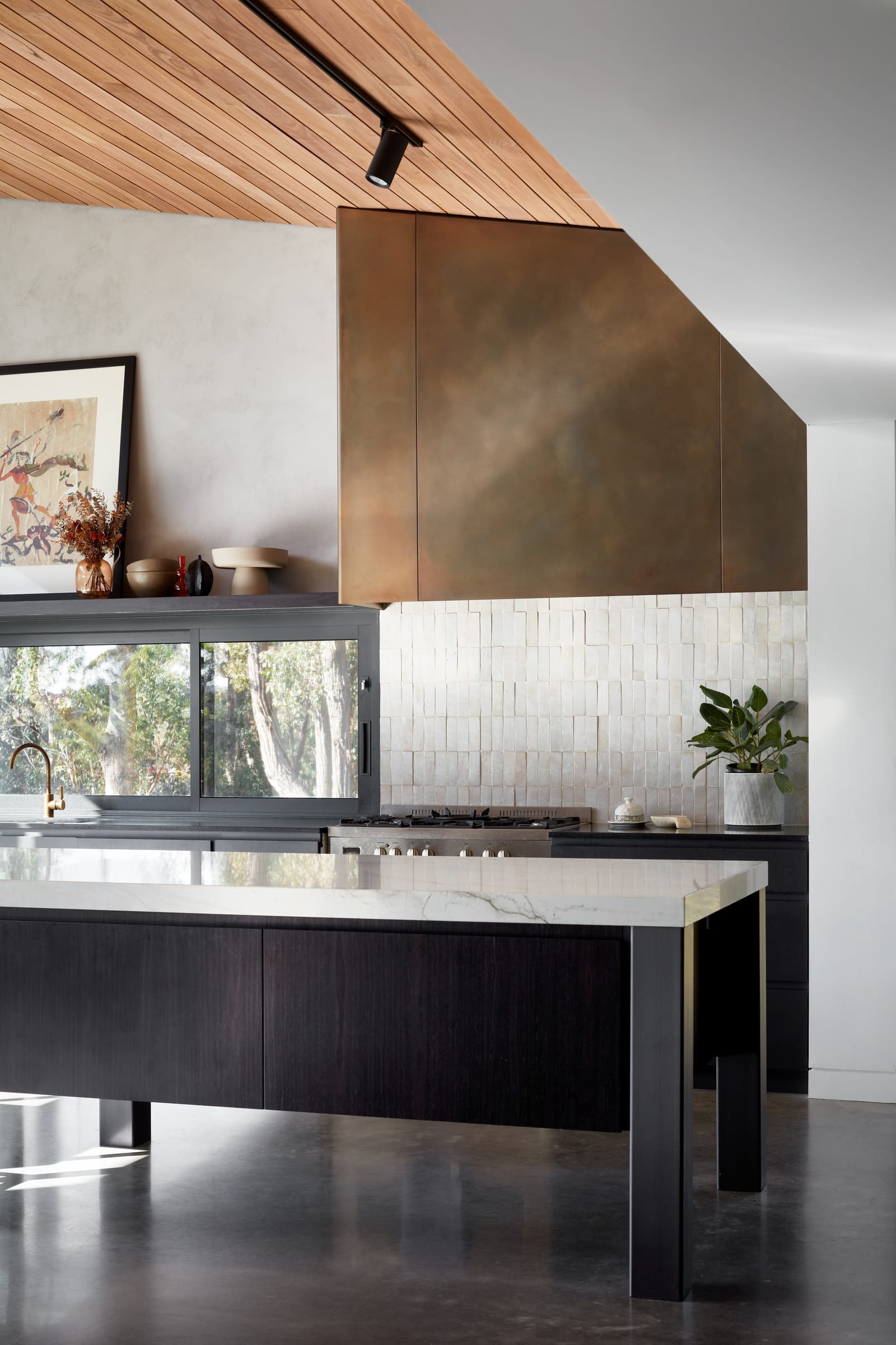 House Woodland by AO Design Studio. The image shows a modern kitchen in House Woodland by AD Design Studio, characterized by its clean lines and a mix of materials. The prominent feature is a large copper hood over the island, contrasting with the white subway tile backsplash. The kitchen has dark countertops and light wooden cabinetry, creating a warm yet contemporary look. The space is well-lit with natural light from the windows.