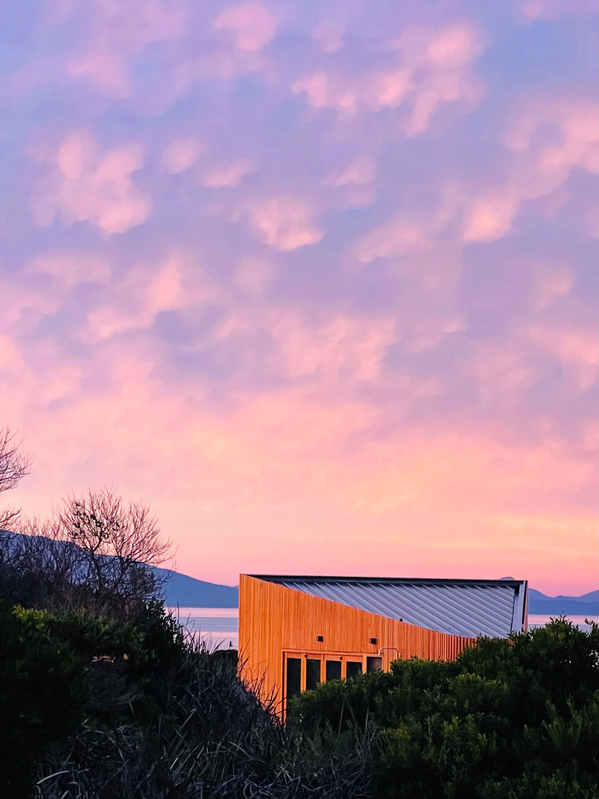 Studio Tasmania in Dolphin Sands, Tasmania. The studio captured at either dawn or dusk, as the sky above transitions through a palette of soft pinks and purples. The silhouette of the building is framed against the calming pastel sky, reflecting the peaceful isolation of the setting by the ocean's edge.