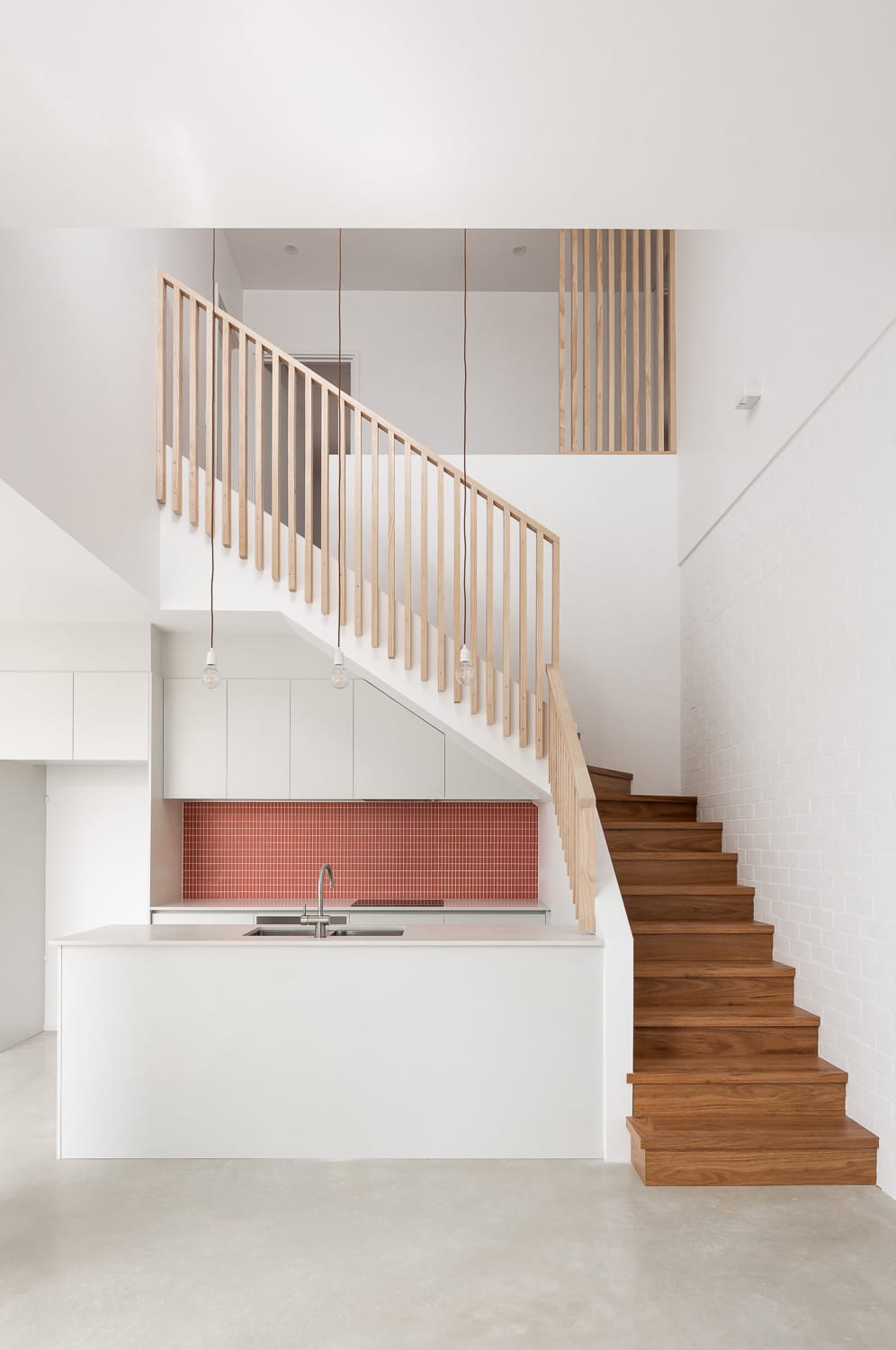 Carrington Street Terraces by MDC Architects.he focus here is on a modern staircase with a minimalist aesthetic. Wooden steps ascend from a white base, leading up to a second level. A simple wooden balustrade lines the staircase, and the walls are white, enhancing the clean, bright feel of the space. In the background, a kitchen area with a red-tiled backsplash is partially visible below the stairs.