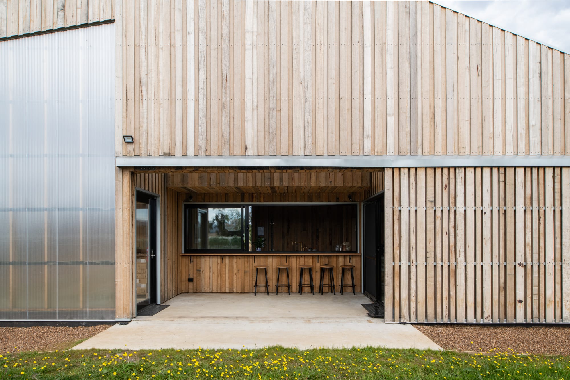 Tasmanian Timber Series: Westella Vineyard.Exterior view of Westella Vineyard's building featuring a weathered timber facade with vertical slats, a large glass entrance, and a corrugated metal roof, blending modern and rustic design elements.