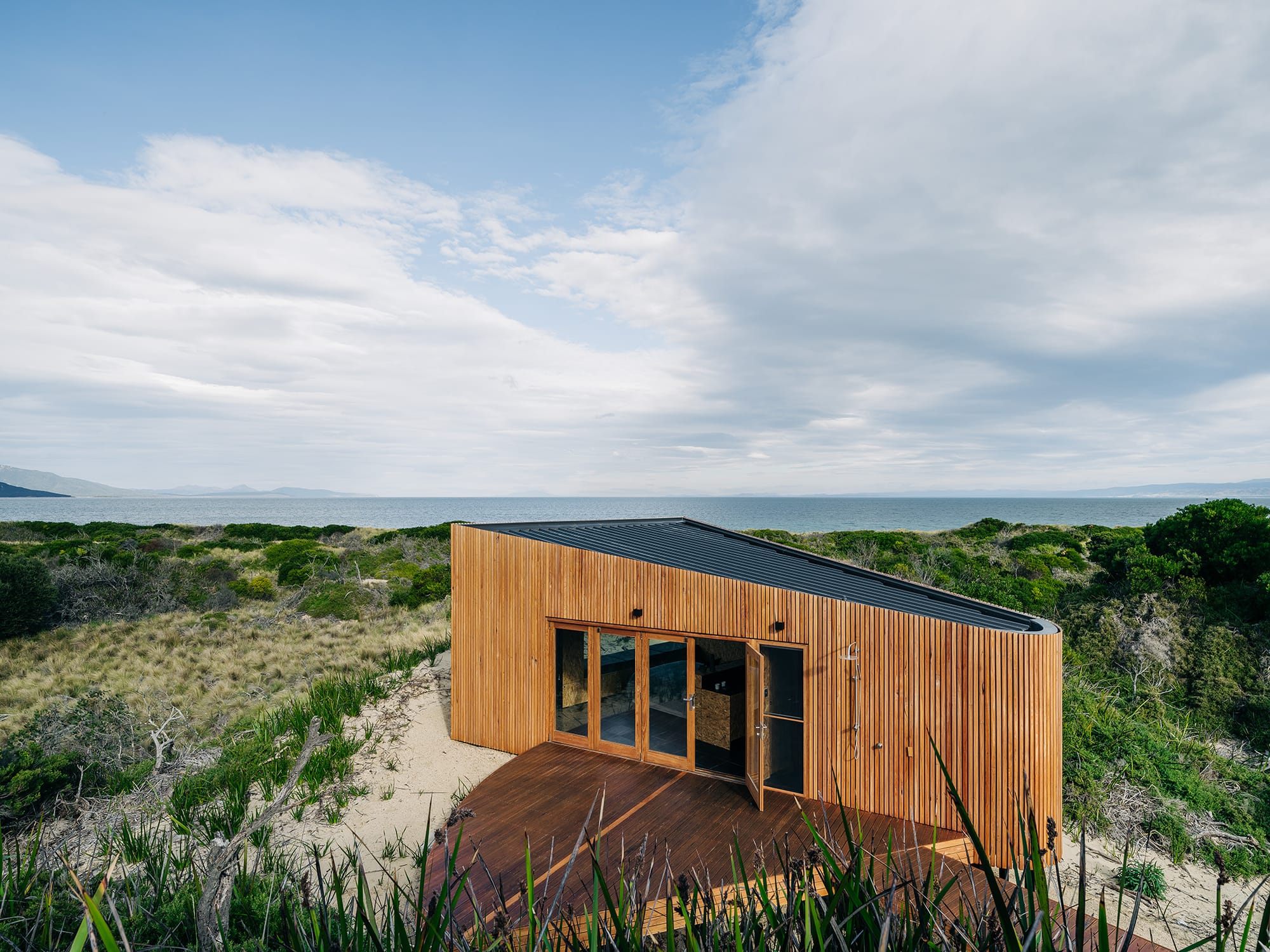 Studio Tasmania in Dolphin Sands, Tasmania. A ground-level perspective showing the studio's wooden exterior and innovative design, which allows it to sit lightly on the landscape. The building's integration with the coastal environment is emphasized by the sandy dune it rests upon, offering a sustainable and immersive connection to the natural setting.