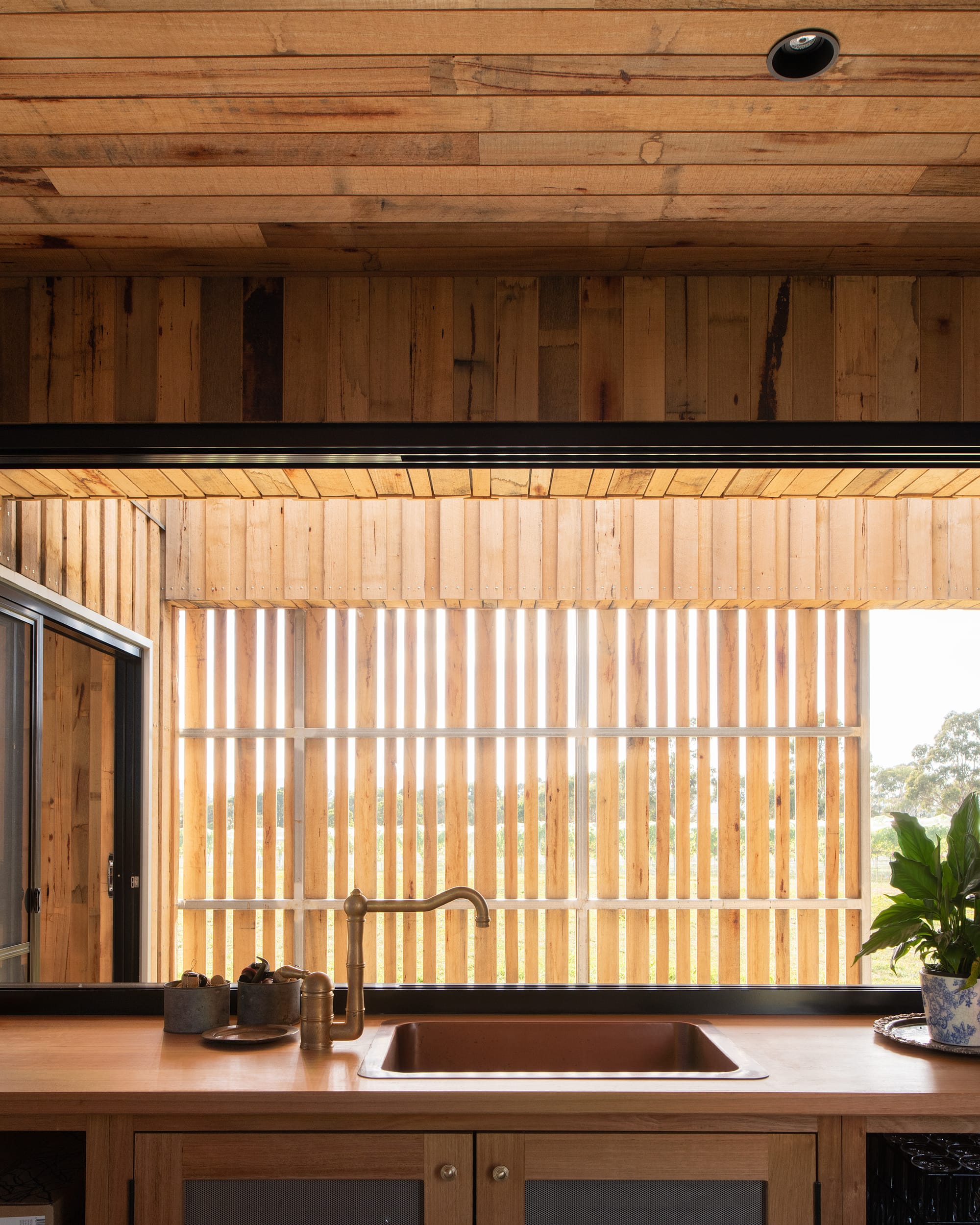 Tasmanian Timber Series: Westella Vineyard.Inside view of a kitchen at Westella Vineyard with natural light filtering through vertical timber slats, highlighting the wooden interior and a copper kitchen sink set within a wooden countertop.