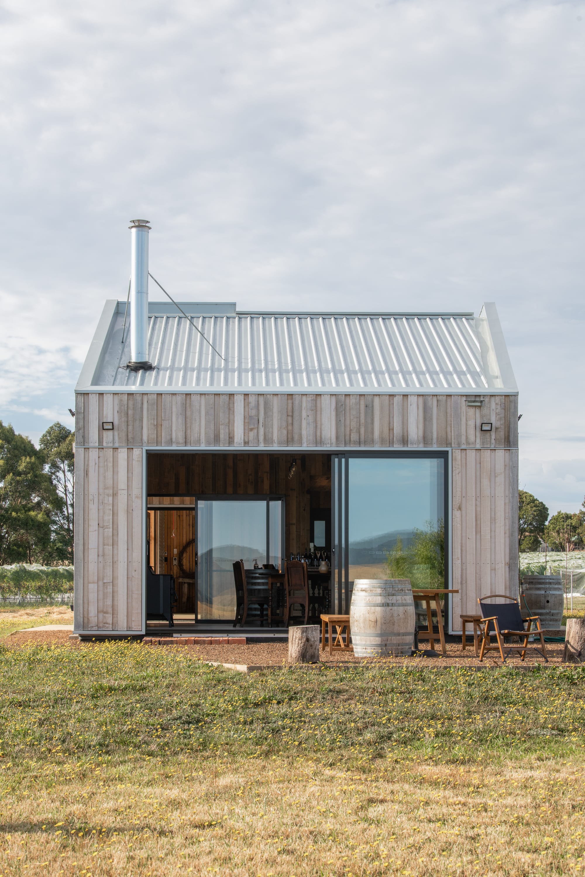 Tasmanian Timber Series: Westella Vineyard.The façade of the winery, featuring large glass doors flanked by timber walls, with a stainless steel chimney and outdoor seating that invites visitors to enjoy the vineyard's serene environment.