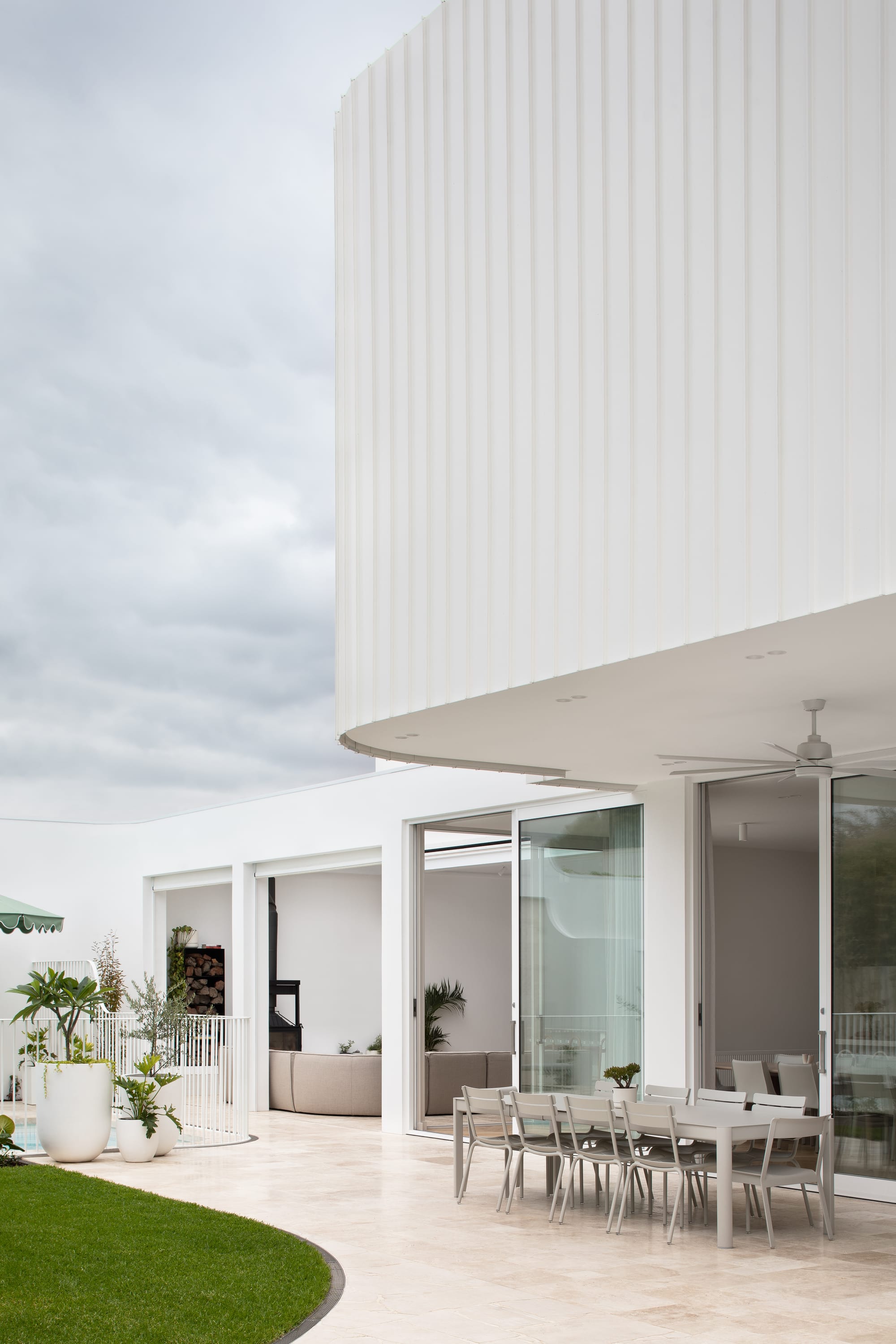 The Grove by Taouk Architects.A modern outdoor dining area adjacent to a minimalist white house with a distinctive vertical profile. The patio area is furnished with a long dining table and chairs, set up for an alfresco dining experience. The house features expansive glass doors that open to seamlessly connect the interior with the exterior, and the landscaped garden adds a touch of greenery to the scene.