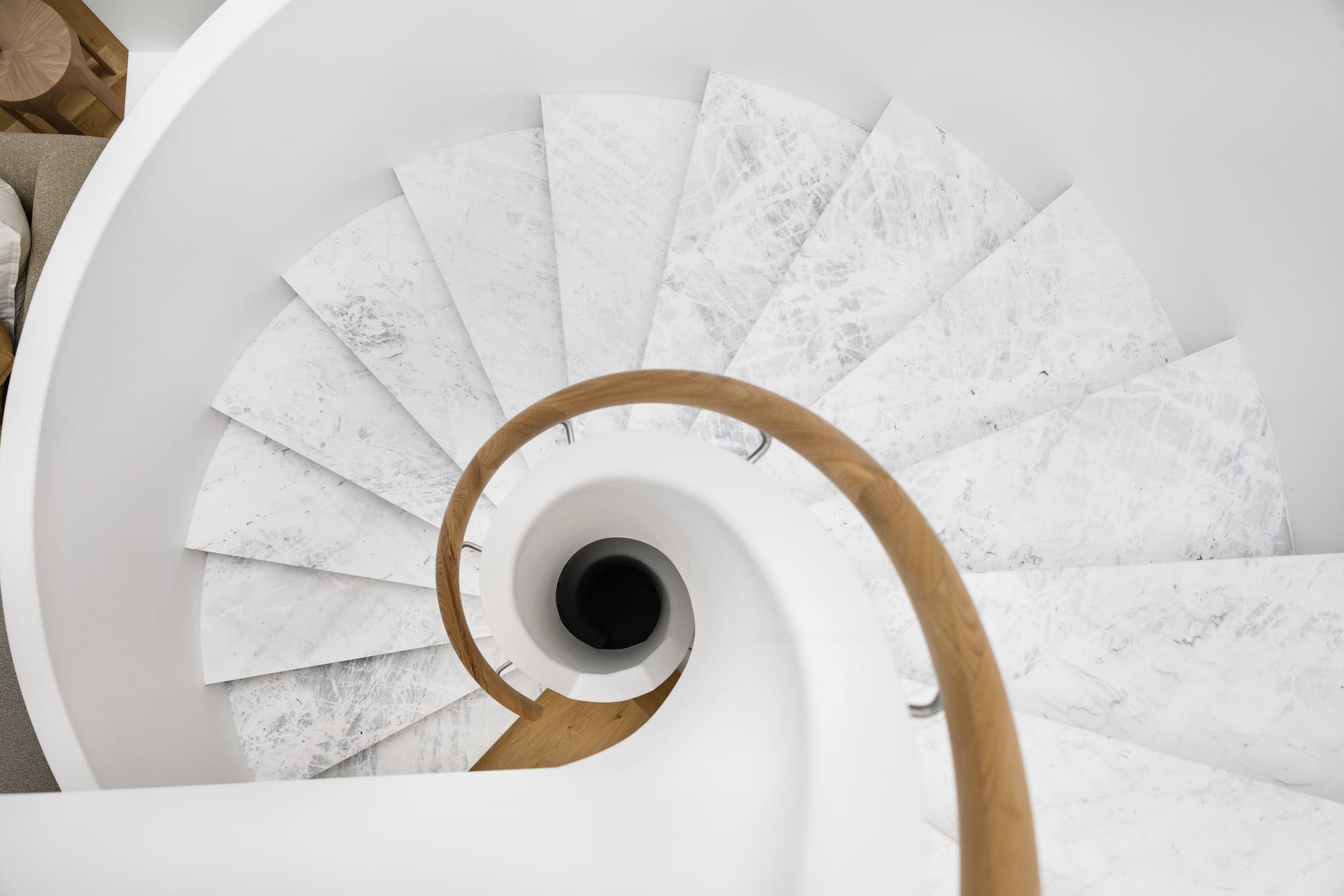 The Grove by Taouk Architects.An elegant view from the top of a white spiral staircase with a classic design, accentuated by a dark brown handrail that contrasts with the pristine white steps. The photo captures the graceful curve of the stairs as they descend, emphasizing the architectural beauty and simplicity of the design.