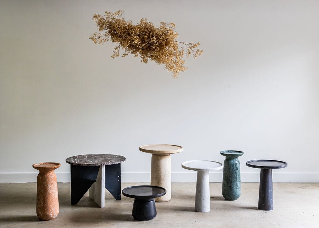 Noma Co Studio. Copyright of Noma Co Studio. Arrangement of different round, marble end tables in a range of beige, orange, white and blue tones. Concrete floor, white walls.
