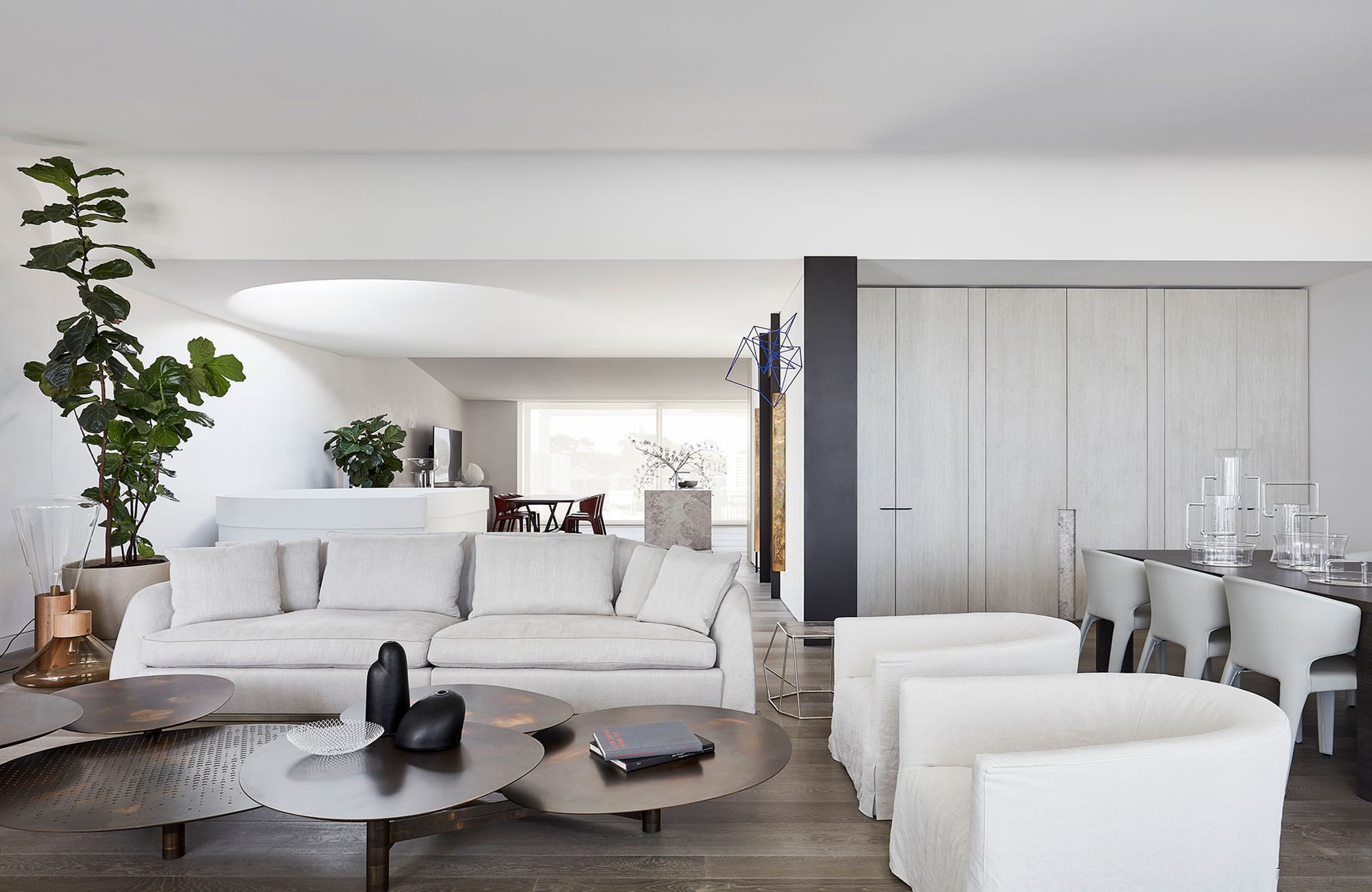 Arc Side by Jolson. Photography by Lucas Allen. Landscape image of residential living space. Tall white walls and ceilings, American Oak timber floors. Plush white couches. Neutral cabinetry along wall. Contemporary, organic coffee table. 