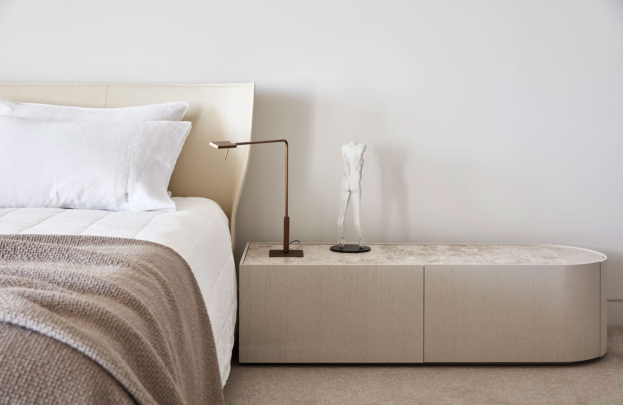 Arc Side by Jolson. Photography by Lucas Allen. Close up landscape shot of residential bedroom. White walls and carpeted floors in neutral, warm tone shade. Long, organically curved bedside table in light timber finish. White bed linen with brown throw rug. 