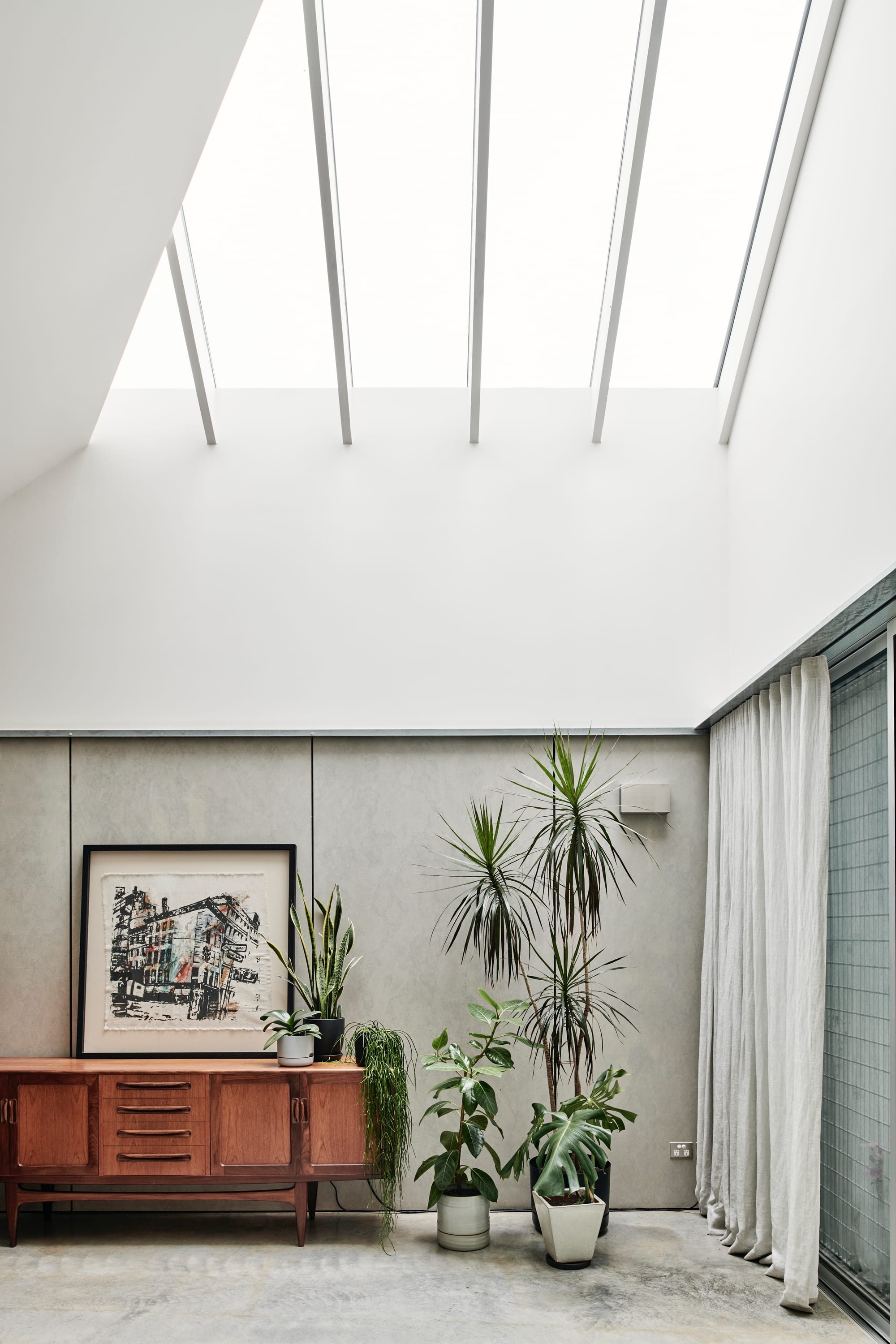 That Old Chestnut by FIGR. Architecture & Design. Photography by Tom Blachford. Residential living room space with concrete floors, cocnrete-finish walls and tall, pitched ceilings with skylights. Space furnished with timber sideboard and plants. 