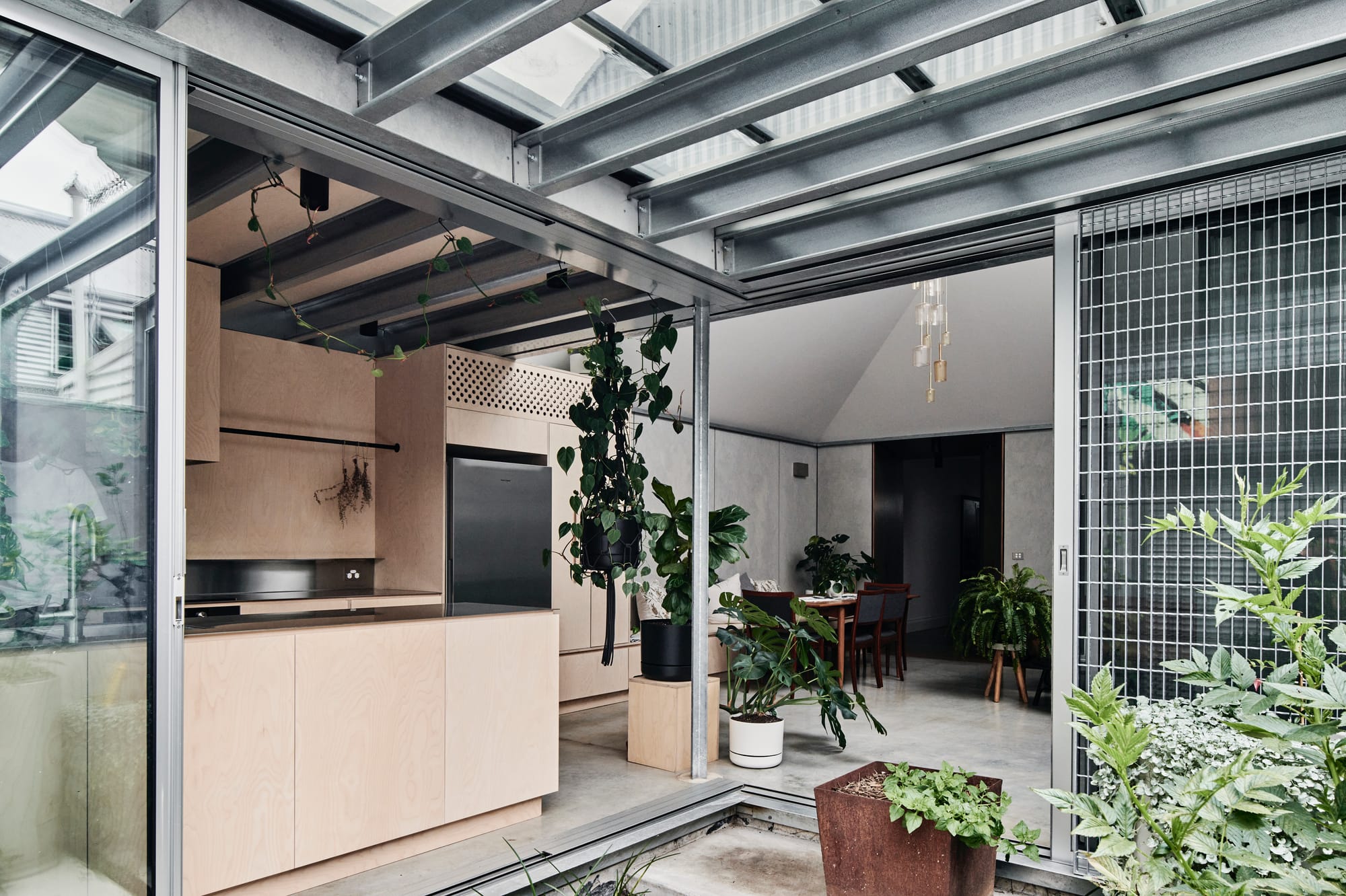 That Old Chestnut by FIGR. Architecture & Design. Photography by Tom Blachford. Landscape image of industrial-style kitchen and living space inside, and courtyard outside. Concrete floors inside, exposed steel beams outside. Plywood kitchen cabinetry. 