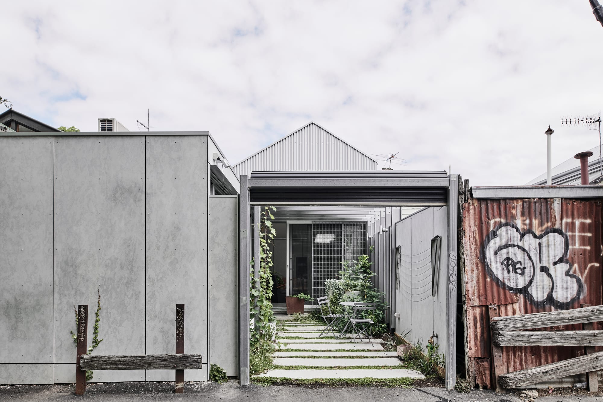 That Old Chestnut by FIGR. Architecture & Design. Photography by Tom Blachford.  Front facade of industrial-style residence. Exposed concrete wall to left of image, exposed and rusted steel to right. Green courtyard of home visible in centre of image. 