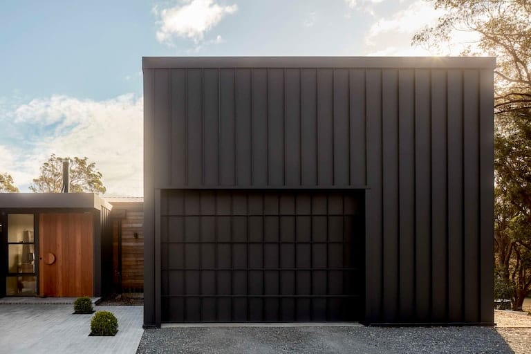 House Woodlands by AO Design Studio. Photography by Luc Remond. Wide angle landscpe image of contemporary external house facade. House is square and panelled in black finish. Modern black garage door in left hand corner of facde. Remainder of house can be seen to left of main building. Large timber door with round handle. 