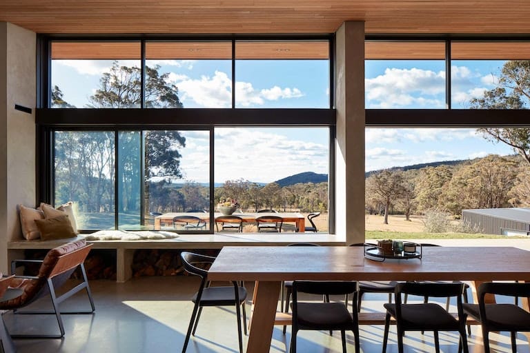 House Woodlands by AO Design Studio. Photography by Luc Remond. Interior of contemporary architectural residential home. Floor to ceiling black framed windows look onto rural landscape. Polished concrete floors and timber veneer roof. Long family timber dining table in centre of room with black modern chairs. 