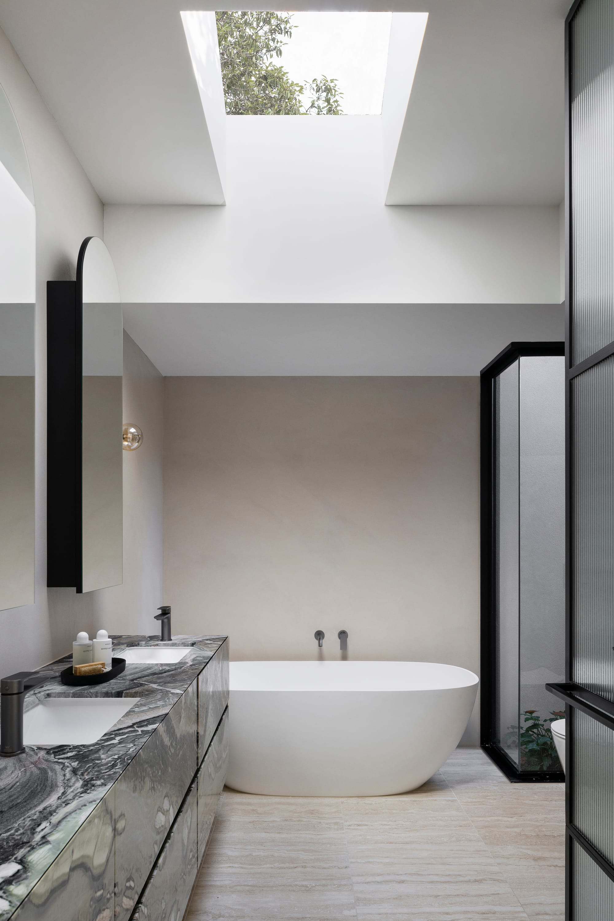 Goldsmith Residence by C.Kairouz Architects. Photography by Spacecraft. Bathroom with beige stone floors, green and black marbled bench and skylight. White freestanding bath. Fluted glass. 
