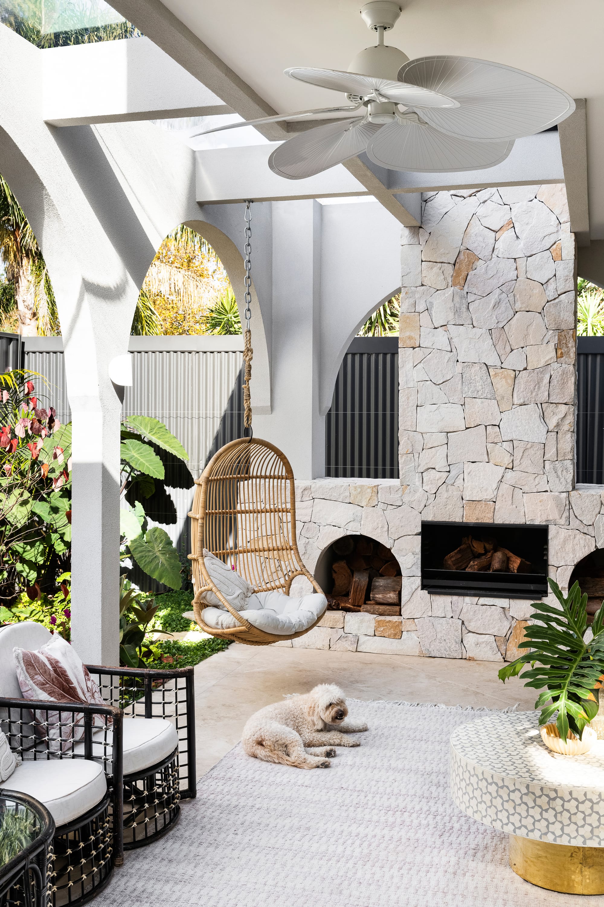 Goldsmith Residence by C.Kairouz Architects. Photography by Spacecraft. Outdoor courtyard with large stone fireplace and wood storage. Hanging rattan chair. White arches line walls. Dog lays on beige stone floor. 