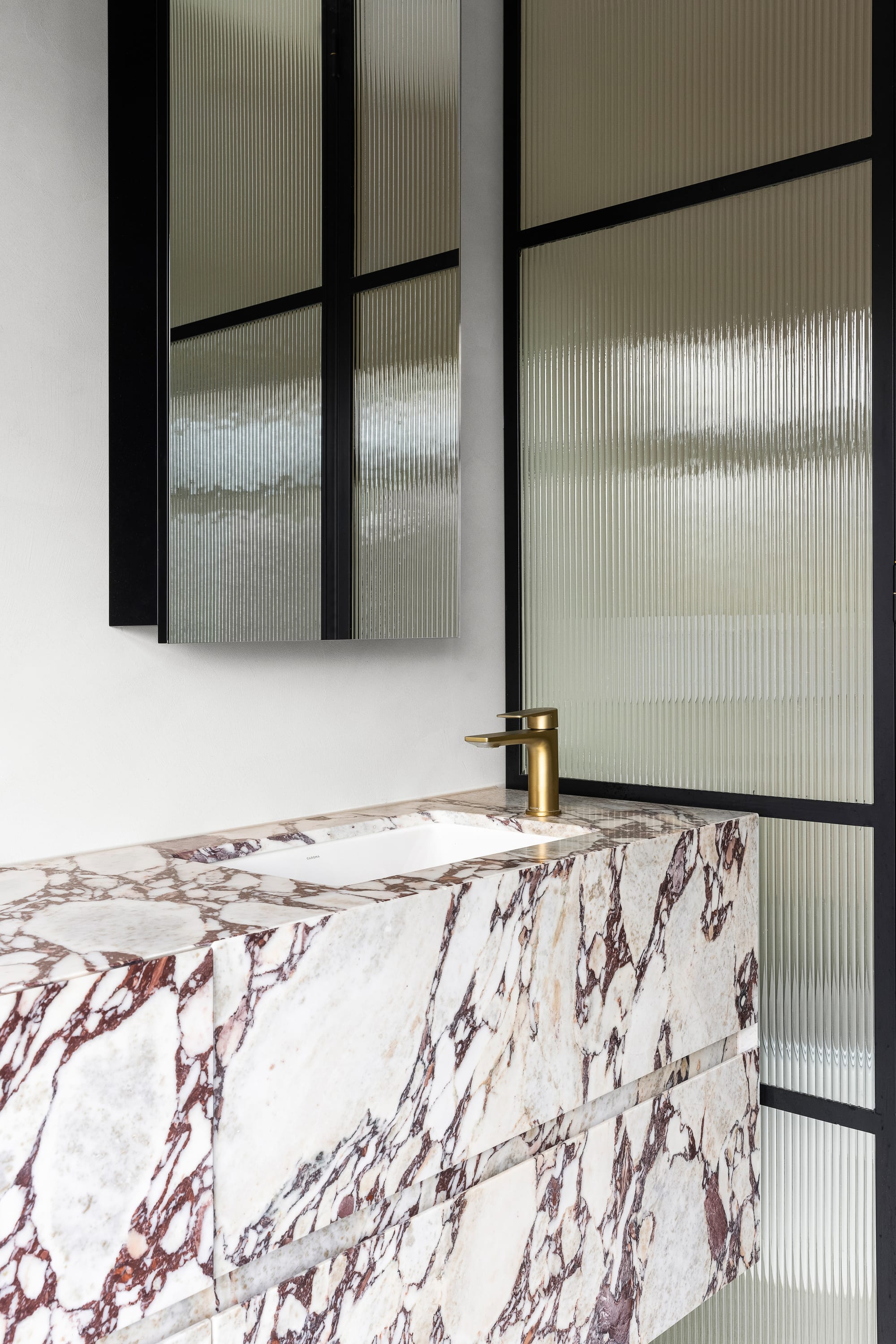 Goldsmith Residence by C.Kairouz Architects. Photography by Spacecraft. White and brown marbled bathroom sink and cabinet with gold tapware. Fluted glass screen with black frame. Black framed mirror above sink. 