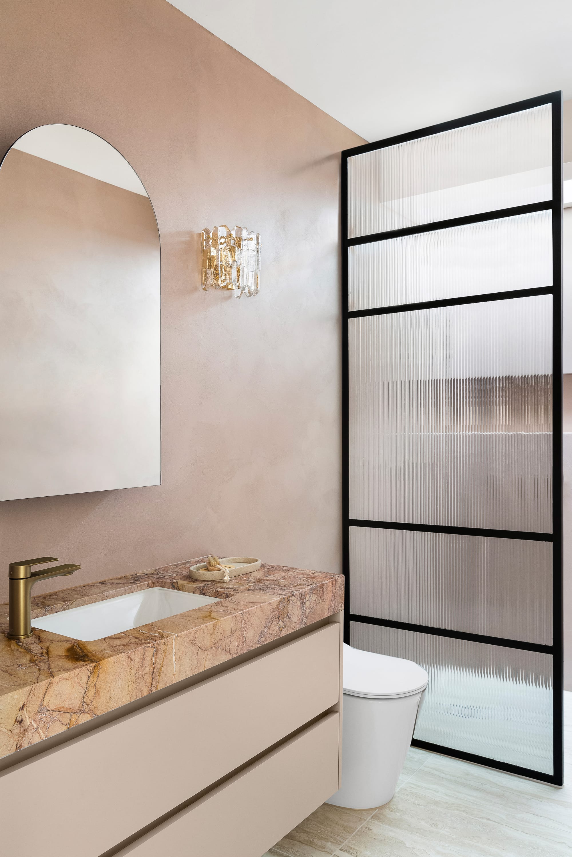 Goldsmith Residence by C.Kairouz Architects. Photography by Spacecraft. Bathroom with peach toned stone benchtop with gold tapware. Black framed fluted glass screen behind white toilet. Pink toned walls behind arched mirror.  