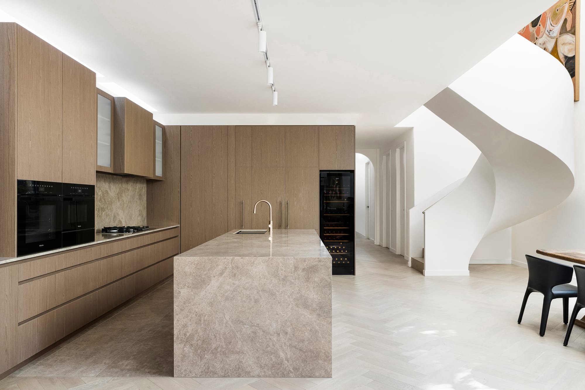 Goldsmith Residence by C.Kairouz Architects. Photography by Spacecraft. Residential kitchen with timber cabinetry and beige stone island bench and benchtops. Pale timber herringbone floors. White curved staircase to right. 
