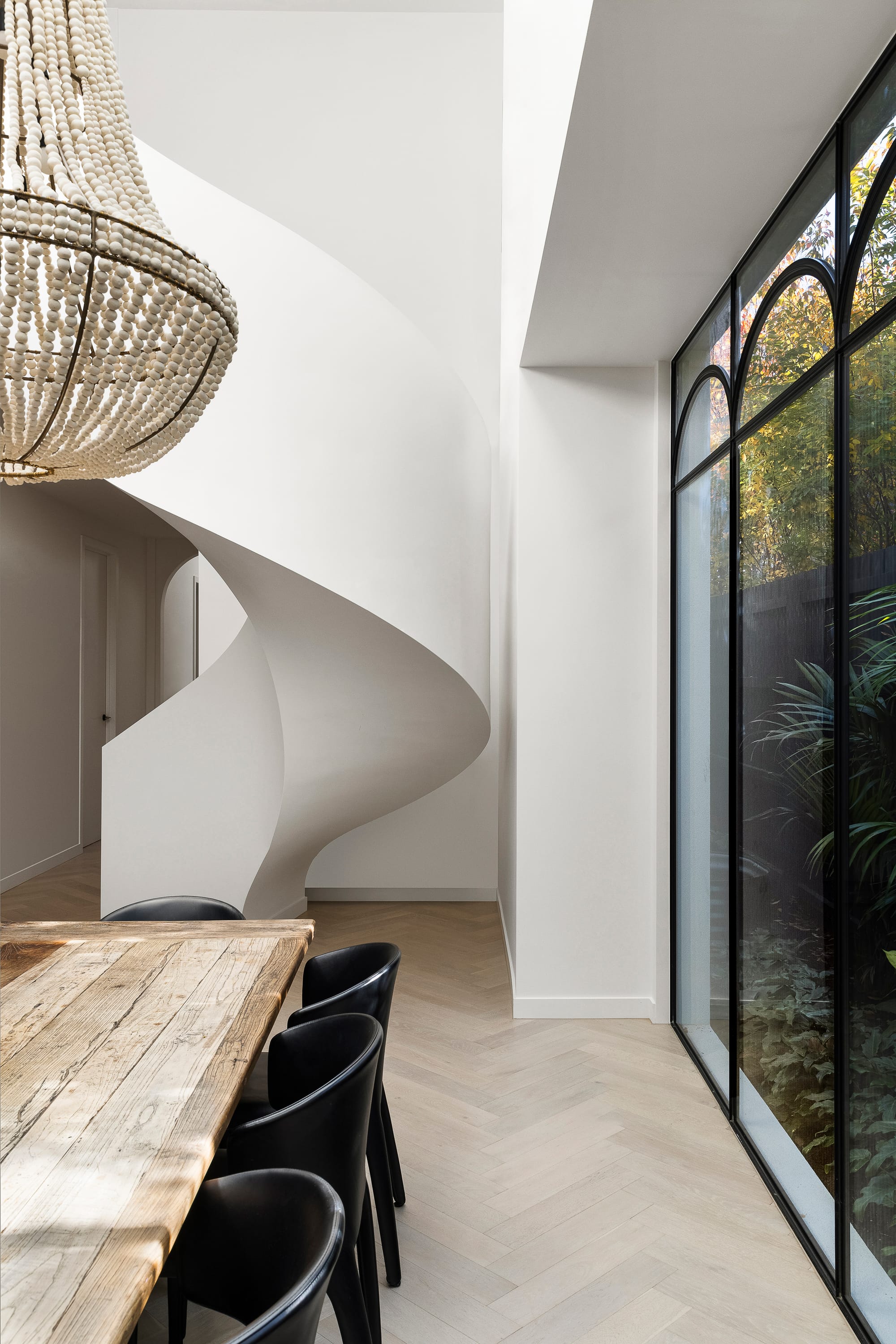 Goldsmith Residence by C.Kairouz Architects. Photography by Spacecraft. Curved staircase behind timber dining table and black chairs. Pale timber herringbone floors. Large beaded pendant light.