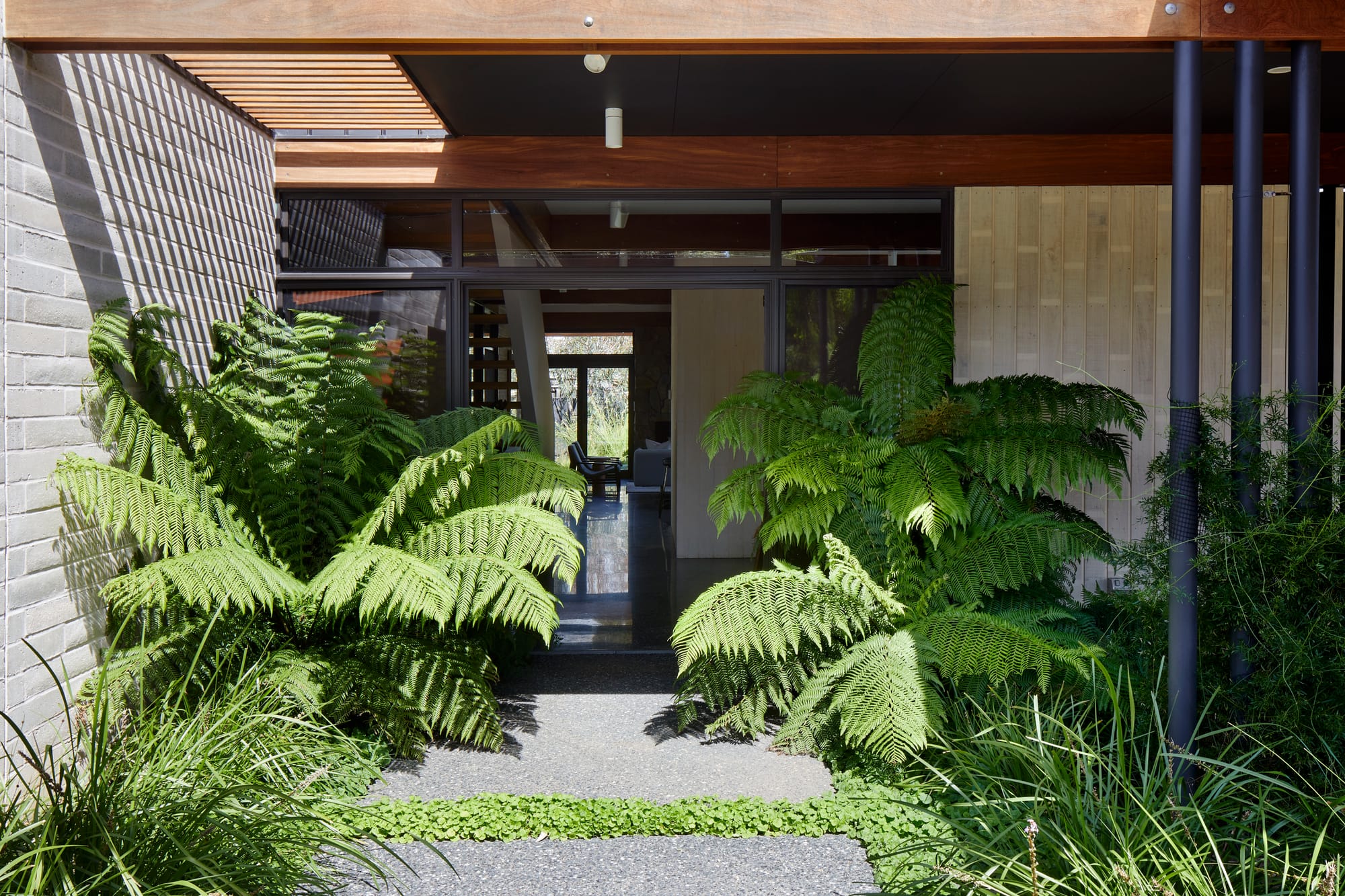 Mt Eliza House by BENT Architecture. Photography by Tatjana Plitt. Residential entryway with concrete pavers and brick wall to left of black framed front door. Large shrubs grow by front door. 