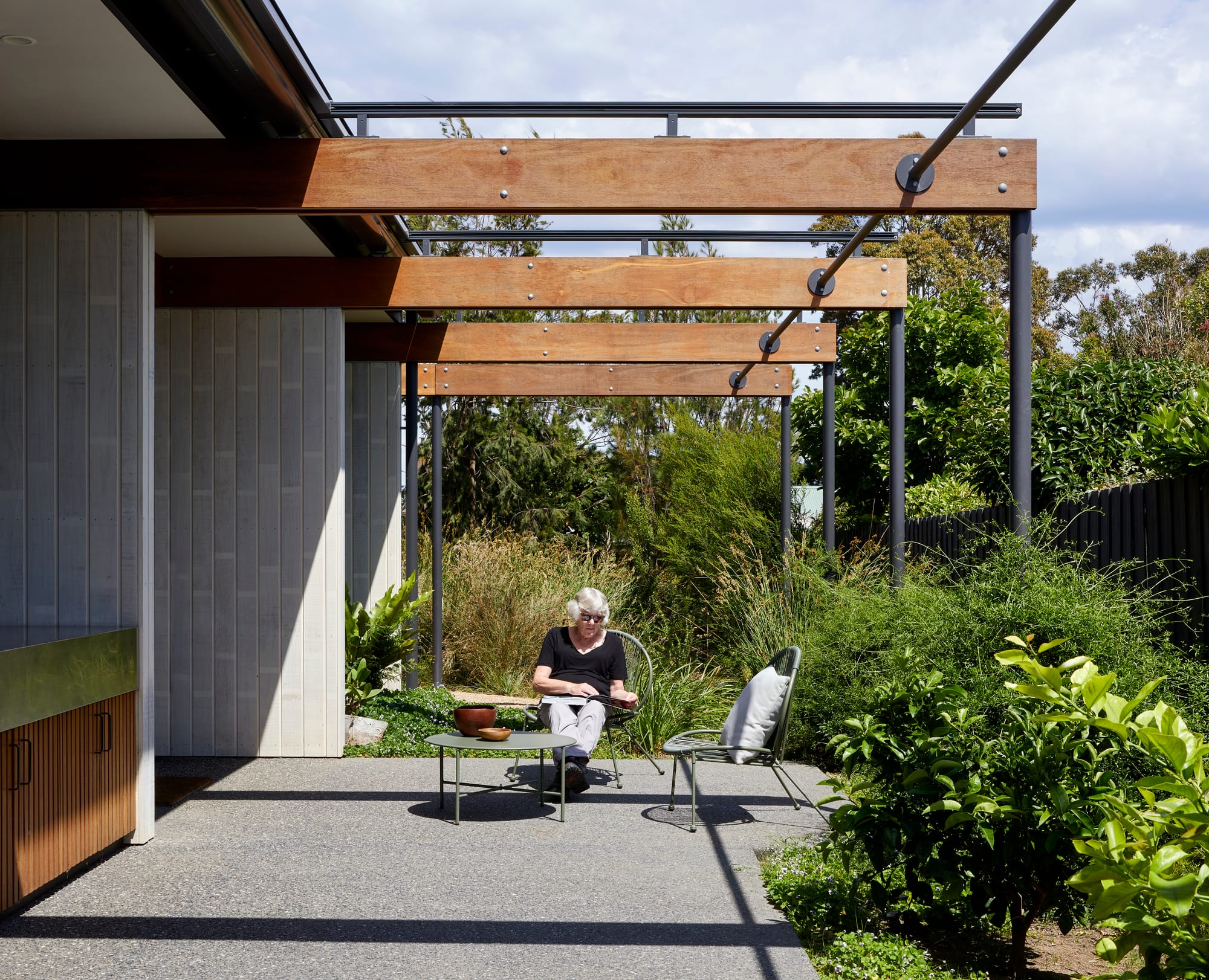 Mt Eliza House by BENT Architecture. Photography by Tatjana Plitt. External patio with concrete floors and brick walls to the left. Exposed timber beams overhand patio floor. Garden grows wildly in background and to right of patio. 