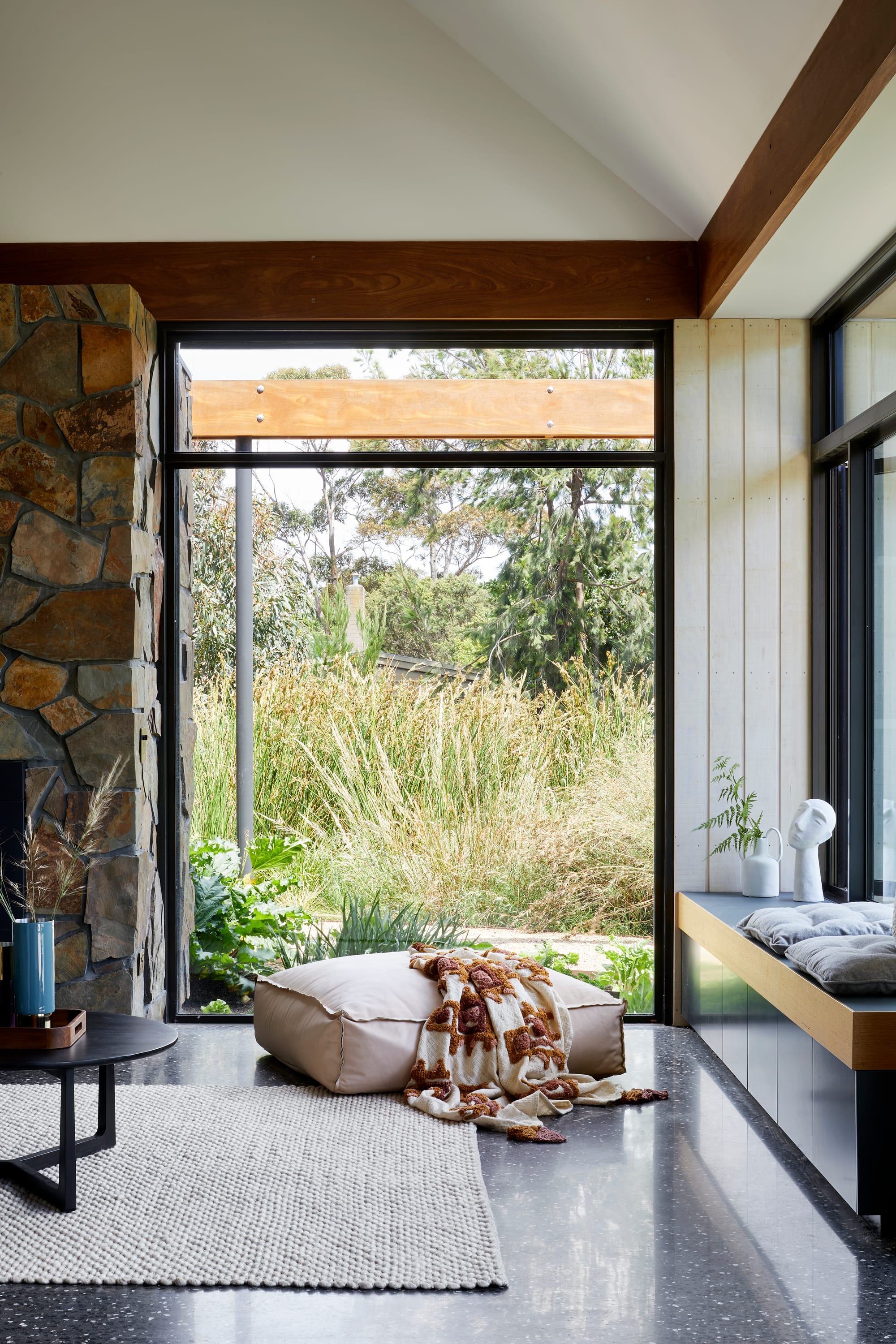 Mt Eliza House by BENT Architecture. Photography by Tatjana Plitt. Living space with floor-to-ceiling black framed windows overlooking garden. Polished concrete floors and cream ottoman on floor. Large stone fireplace to left of image.