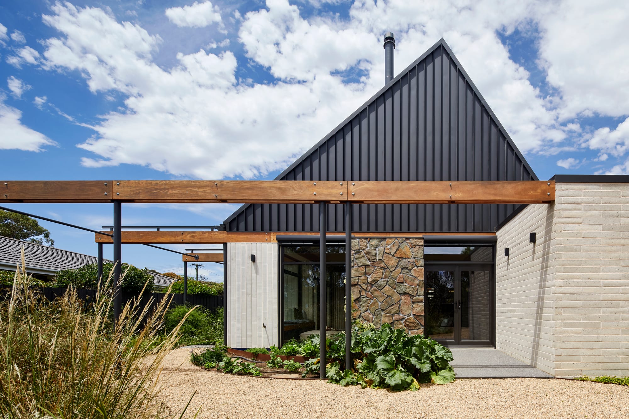 Mt Eliza House by BENT Architecture. Photography by Tatjana Plitt. Exterior rear facade of residential home with light brick walls, black metal roof and feature stone wall. Crushed stone path around house. 
