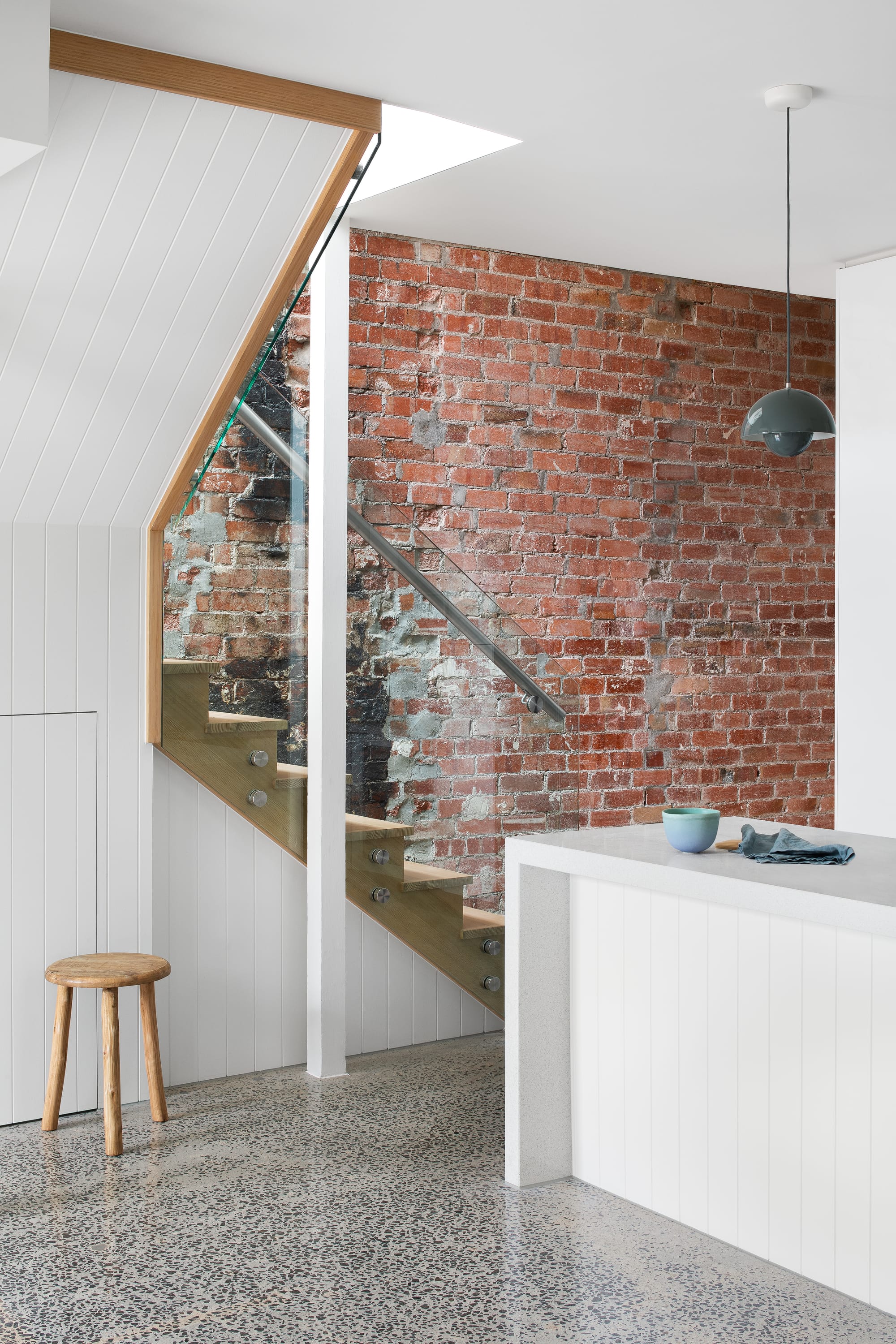 Stead House by Hannaford Design Studio. Photography by Emily Bartlett. Exposed brick wall behind timber staircase with glass balustrade. White clad kitchen island with white countertop. 
