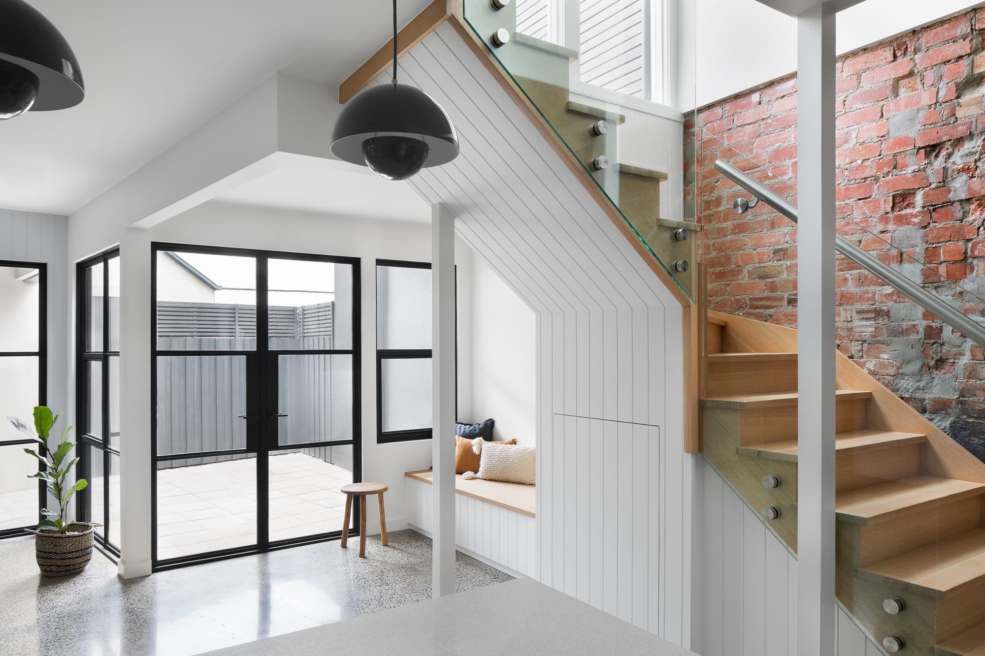 Stead House by Hannaford Design Studio. Photography by Emily Bartlett. Interior of ground floor of residential home with timber staircase to the right and black framed French doors to the left. Polished concrete floors. Exposed brick wall. 