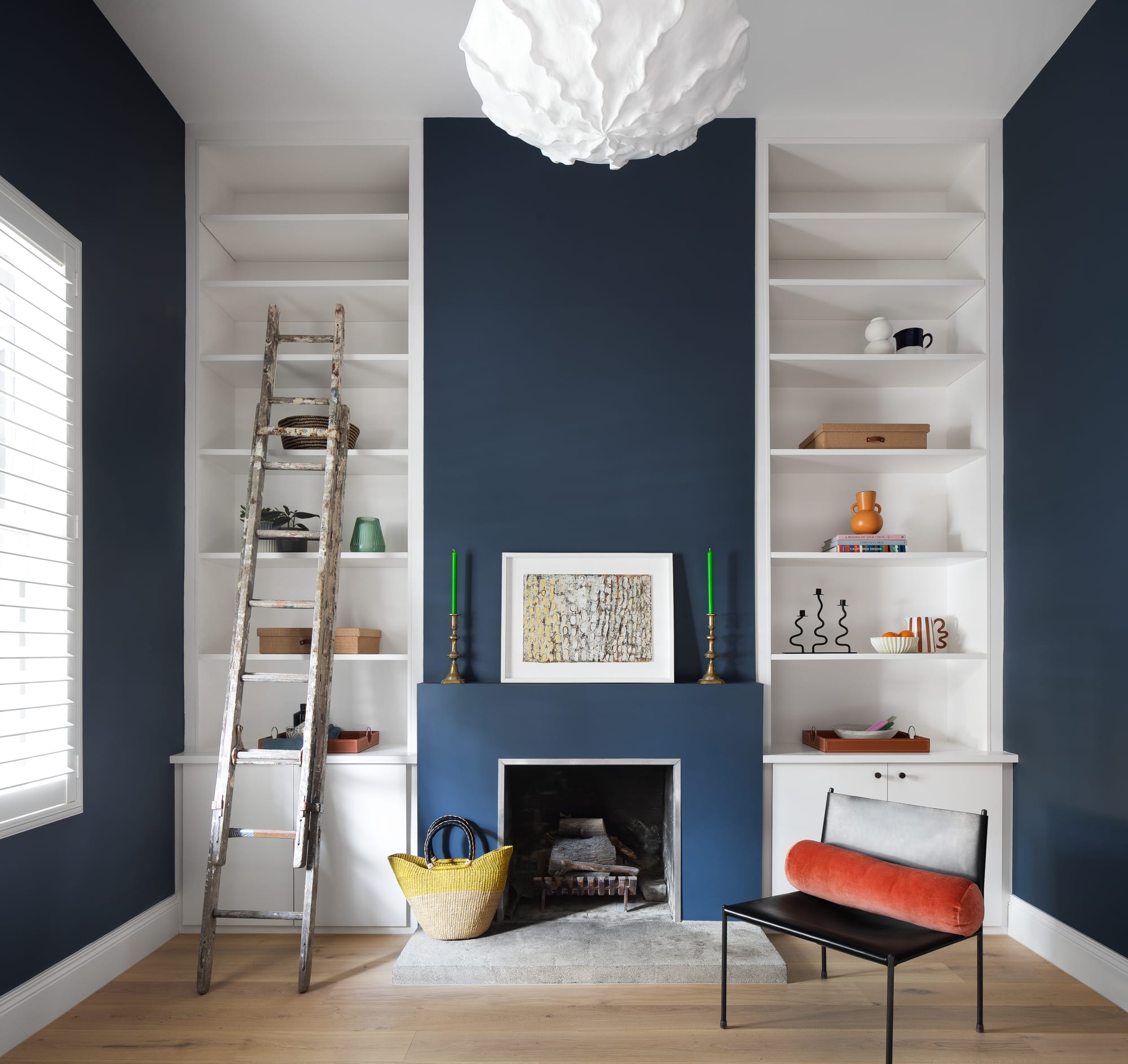 Stead House by Hannaford Design Studio. Photography by Emily Bartlett. Living space with navy feature wall and fireplace. Timber floors and white floor-to-ceiling shelving. Leather chair on right with orange cushion.