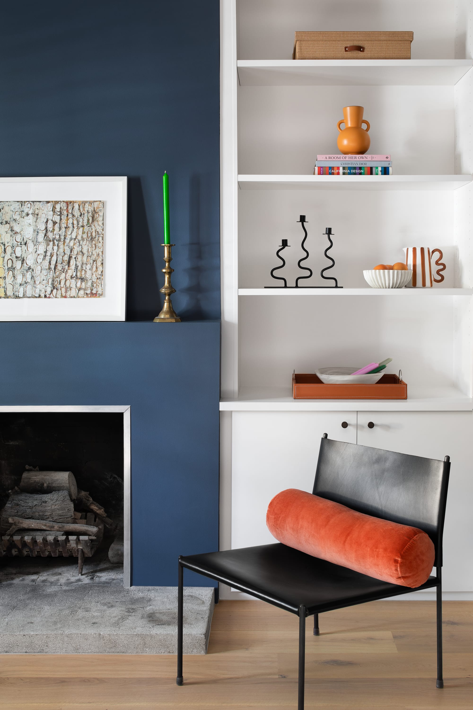 Stead House by Hannaford Design Studio. Photography by Emily Bartlett. Leather chair with orange cushion in front of navy wall and fireplace. White shelves. Timber floors. 