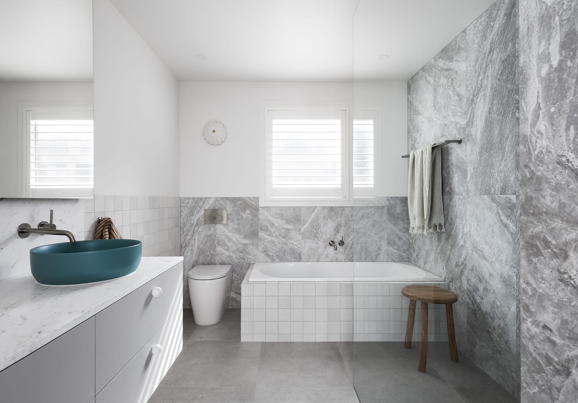 Stead House by Hannaford Design Studio. Photography by Emily Bartlett. Landscape shot of bathroom with grey floor tiles, grey marble wall tiles and white tiled bath. Blue sink on grey cabinets.
