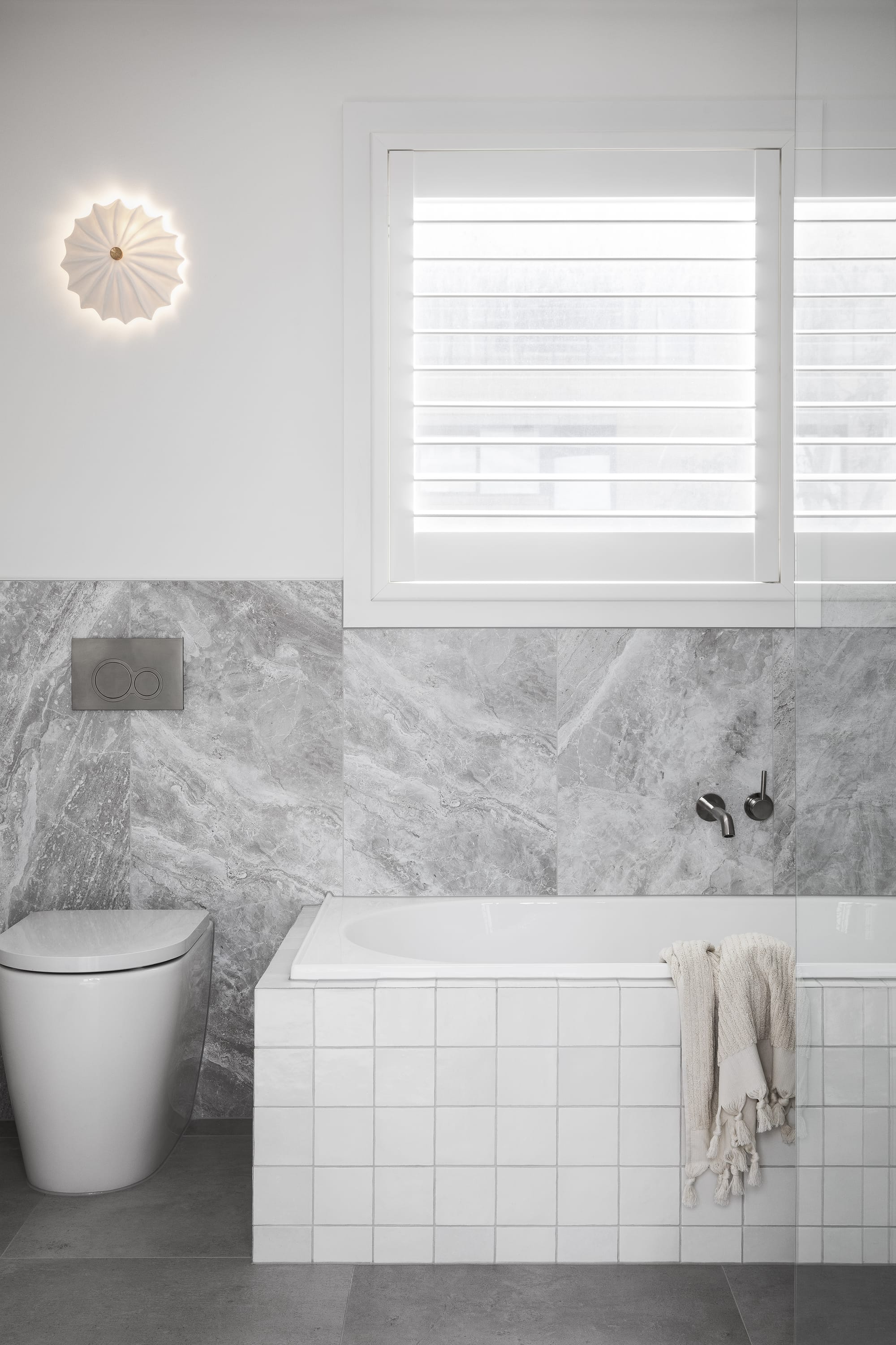 Stead House by Hannaford Design Studio. Photography by Emily Bartlett. Marble tiled bathroom with white bath and toilet. Plantation shutters over the windows. 