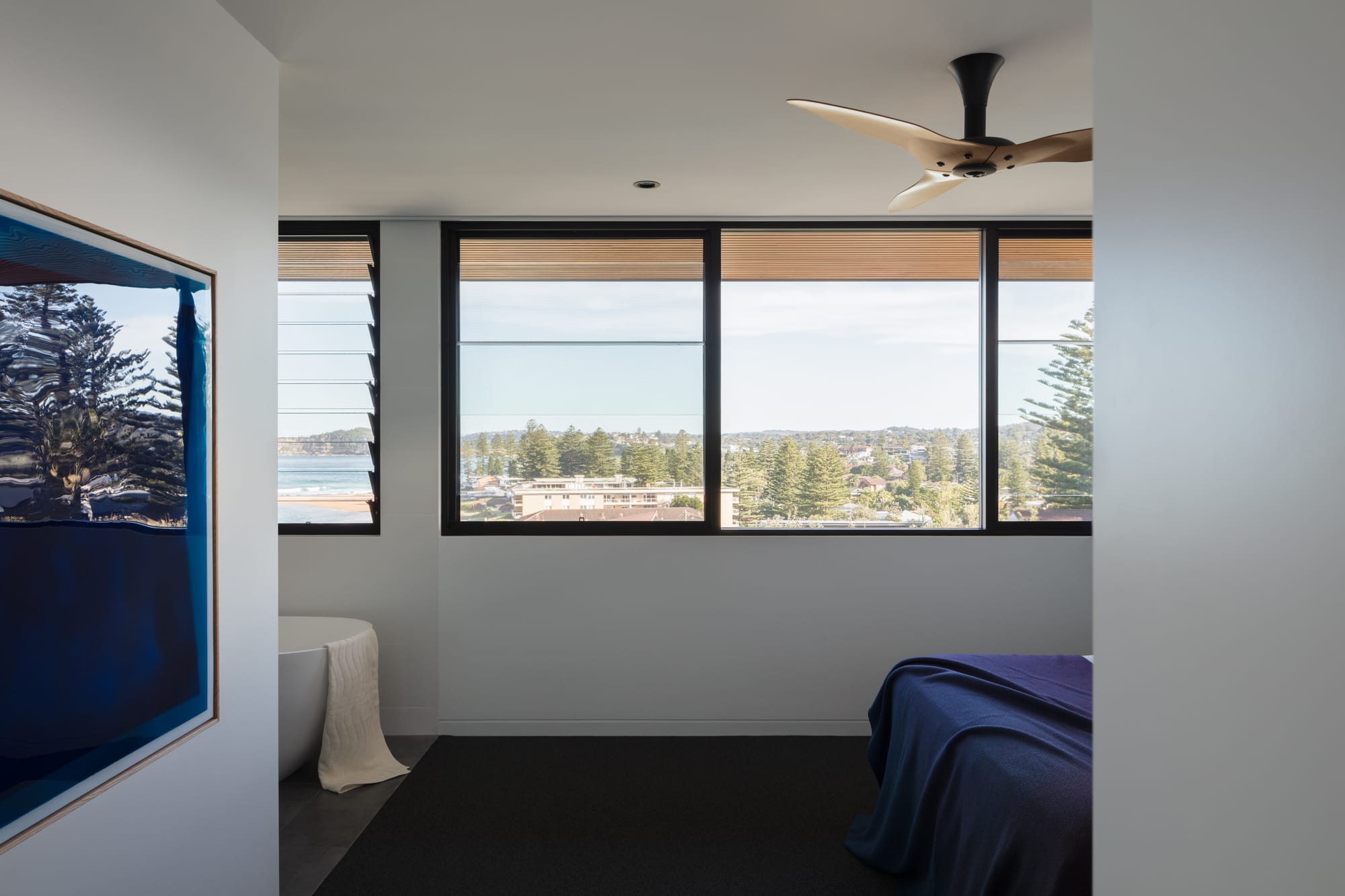 Grandview House by Ian Bennett Design Studio. Photography by Clinton Weaver. Residential bedroom with black framed windows overlooking the beach. Dark carpets, blue bed spread and blue artwork on left wall. 