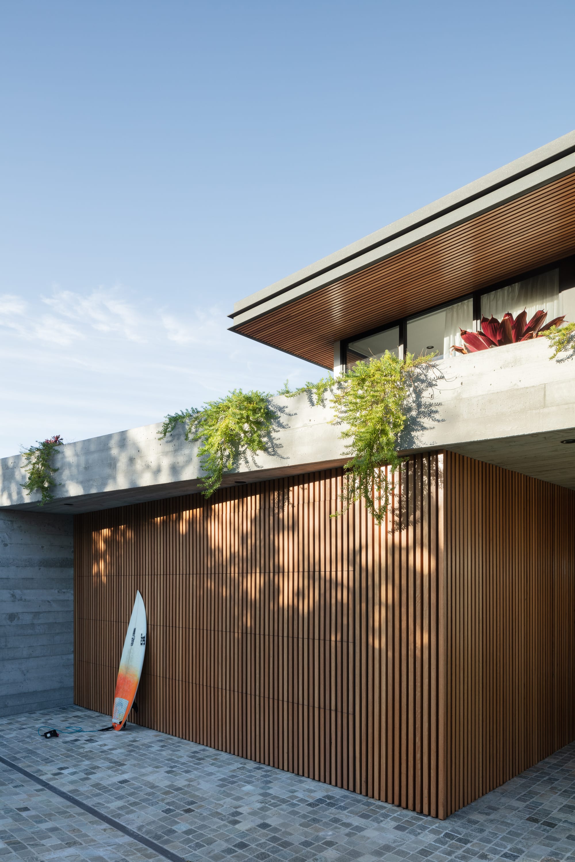 Grandview House by Ian Bennett Design Studio. Photography by Clinton Weaver. Close-up shot of street facade. Grey paved driveway, timber clad garage door and concrete surrounds. Surfboard leaning against timber clad. Plants growing over concrete. 