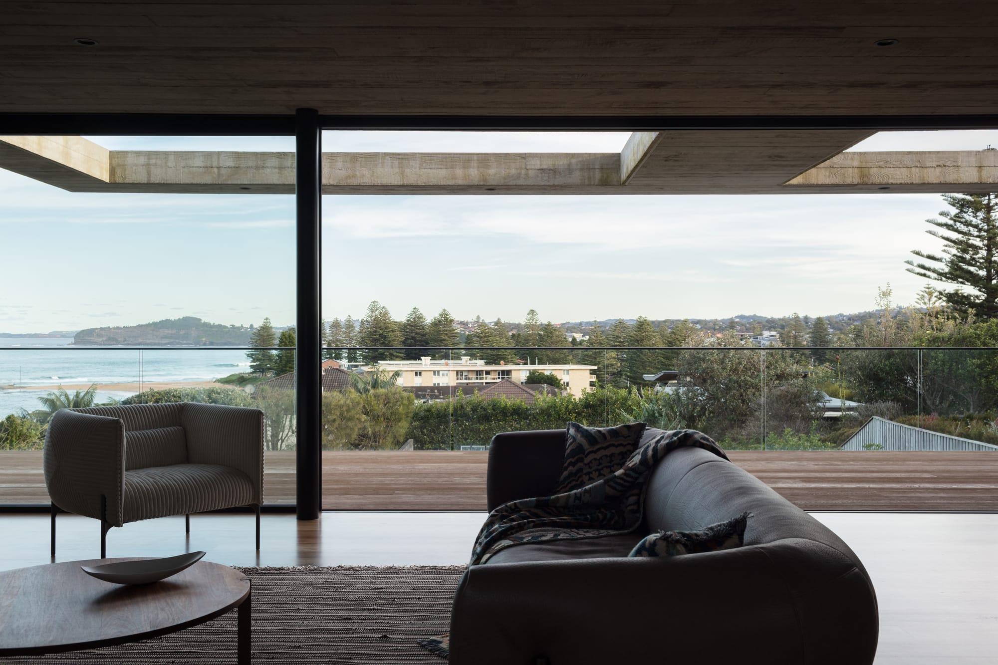 Grandview House by Ian Bennett Design Studio. Photography by Clinton Weaver. Living area with floor-to-ceiling windows opening onto timber balcony. Coastal views in background. 