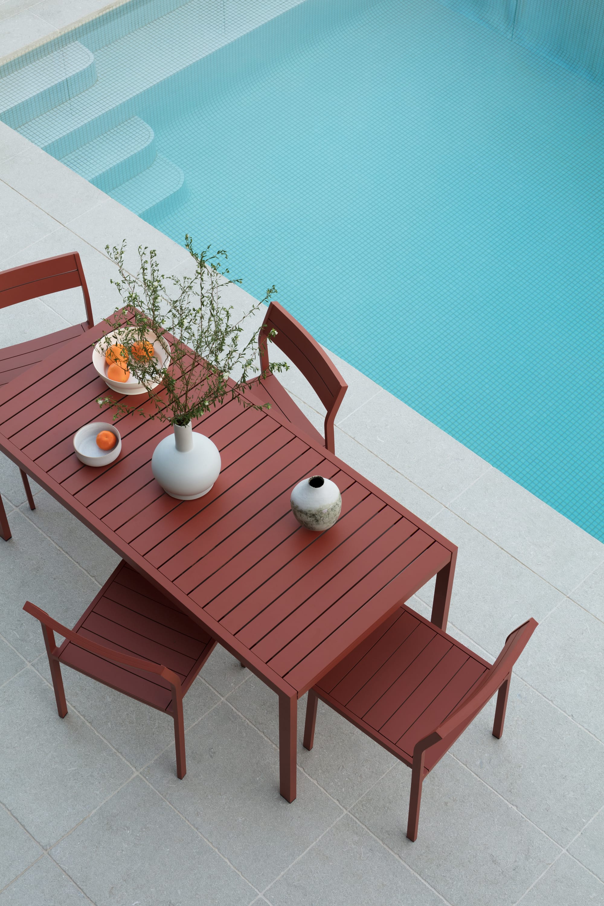 Grandview House by Ian Bennett Design Studio. Photography by Clinton Weaver. Birds eye view of terracotta red coloured outdoor dining set on pale tiles. Tiled pool to right of table. 