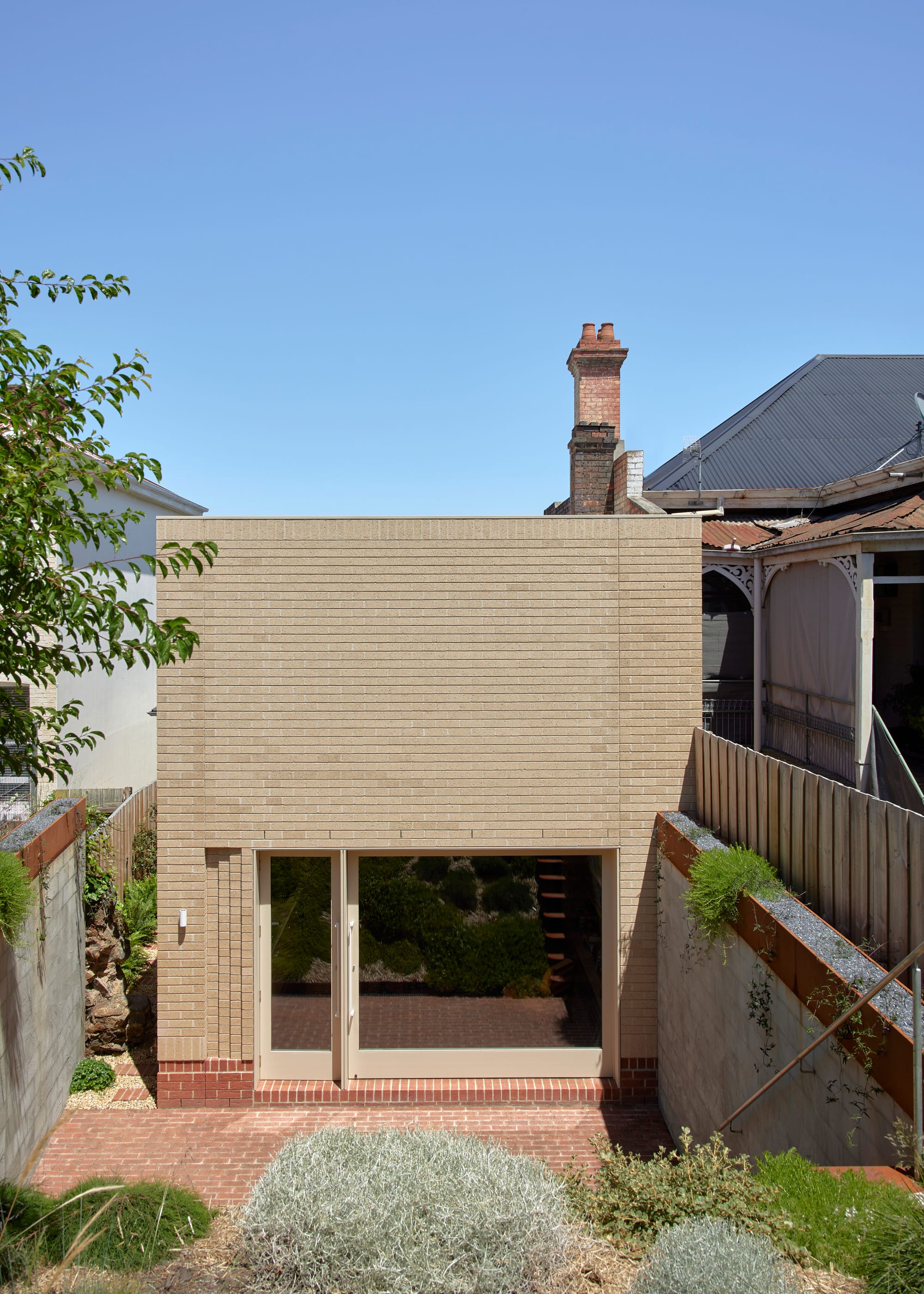 Harriet's House by SO:Architecture. Photography by Sean Fennessy. Rear facade of home. Pale brick single story home with tall concrete-block fences and lush gardens in the foreground.