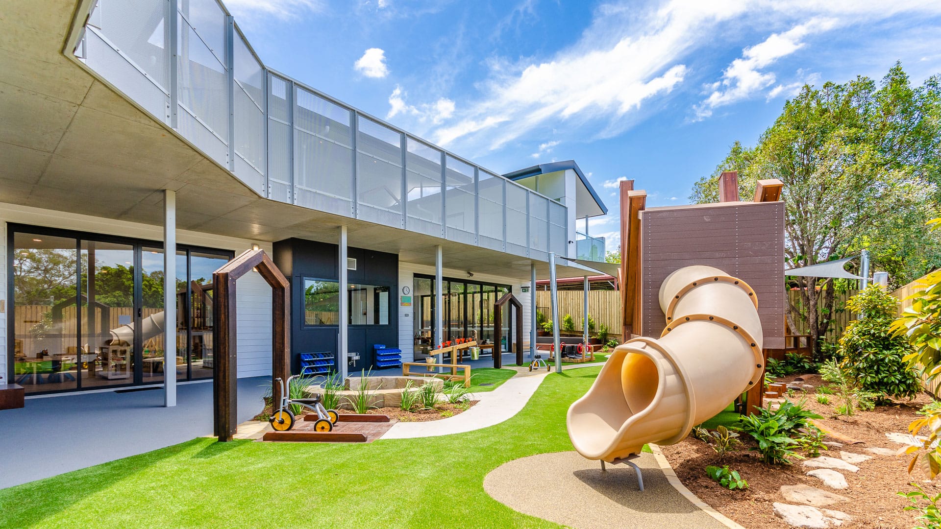 Little & Co. Early Education Childcare Centre by 77 Architecture. Copyright of 77 Architecture. Exterior playground at childcare centre. Multi-level building to left of image. Slide and garden to right. 