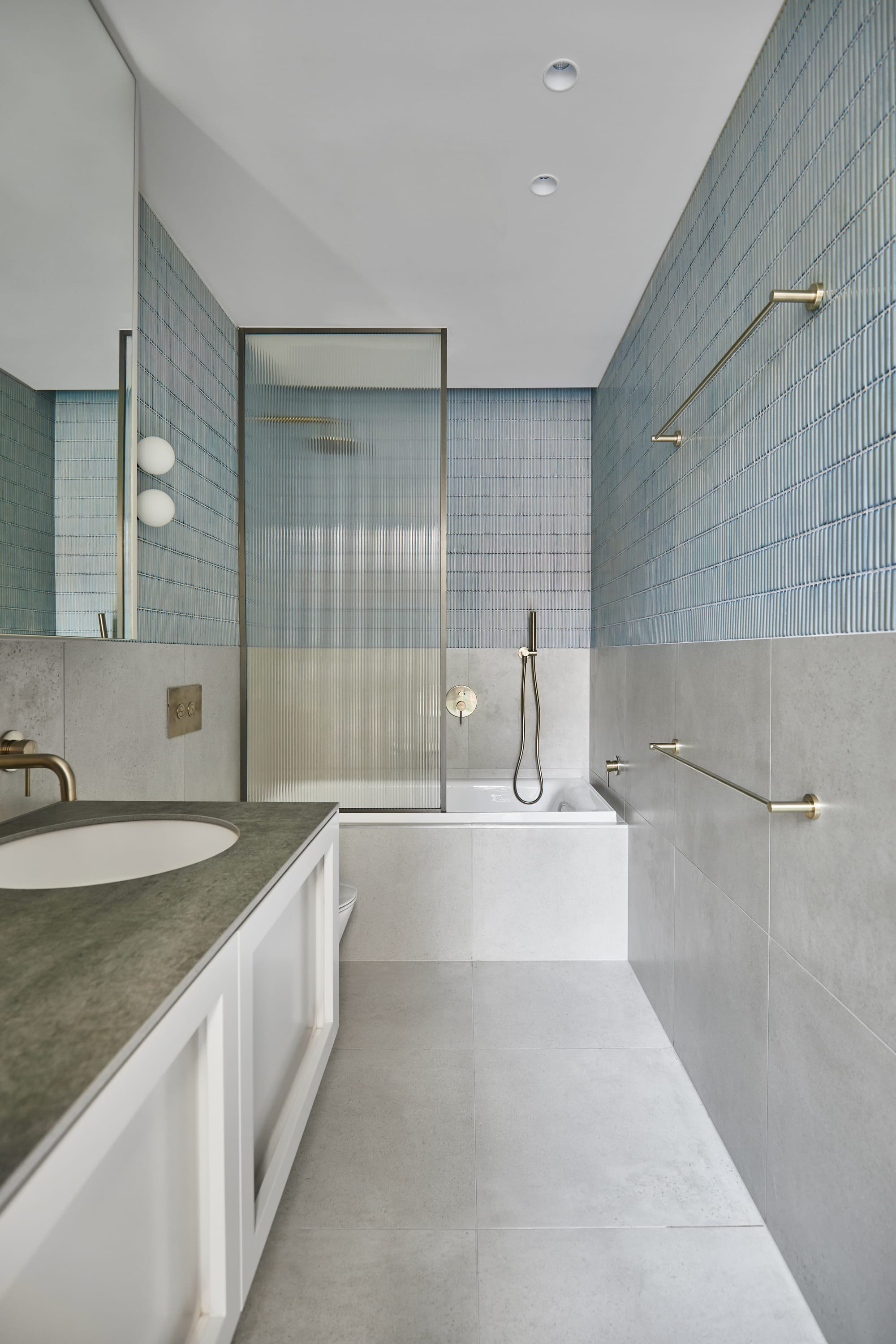 The Full Monty by FYC Architects. Photography by Stephanie Rooney. Bathroom with pale grey floor and wall tiles, blue finger tiles, grey countertop, fluted glass shower screen and white cabinetry. 