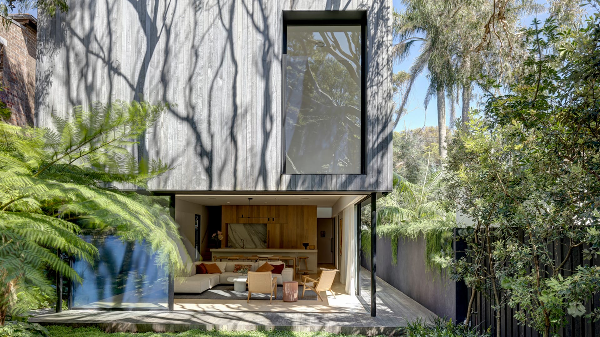 Quarry Box by MCK Architects. Photography by Brett Boardman. Ladnscape image of rear facade of home with large opening on ground floor onto timber deck, lush garden and open plan living. Exterior clad in aged timber with large black framed window on second floor. 