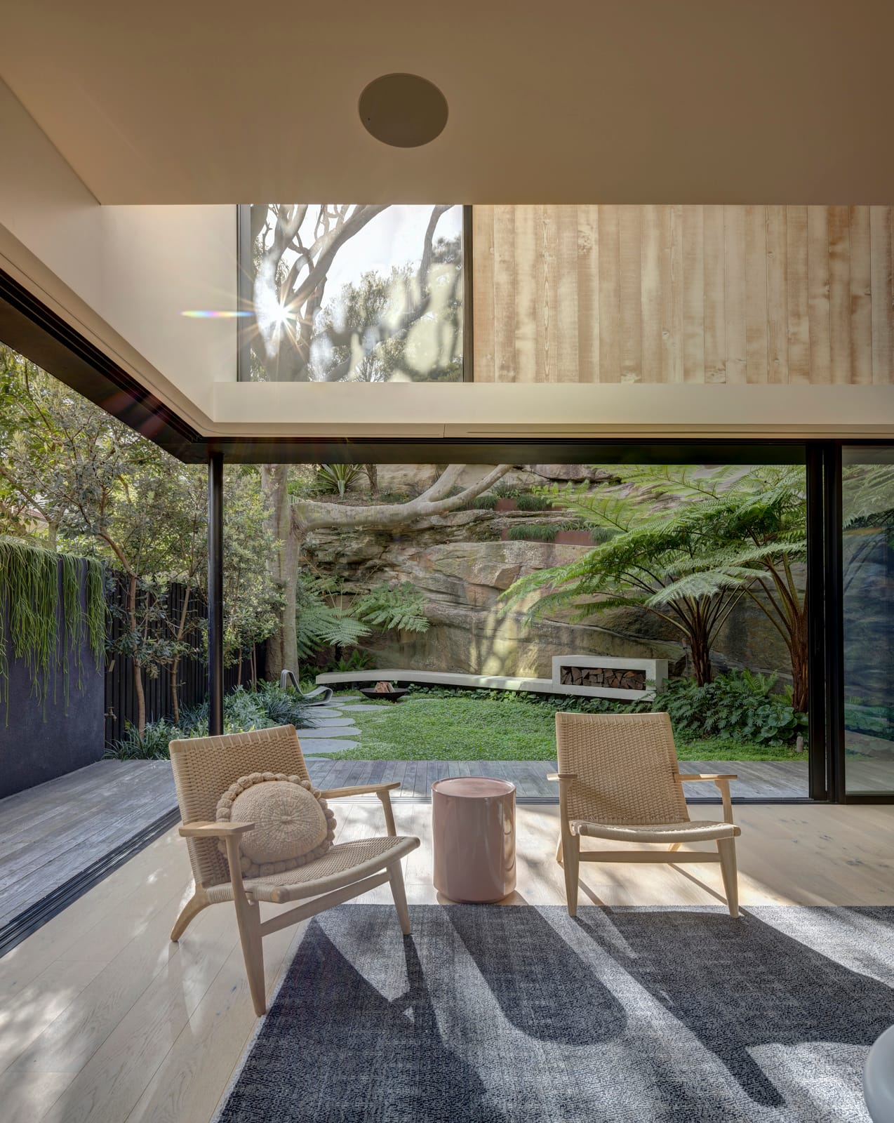 Quarry Box by MCK Architects. Photography by Brett Boardman. Living room with two cane chairs on grey rug. Floor to ceiling windows overlooking backyard behind chairs.