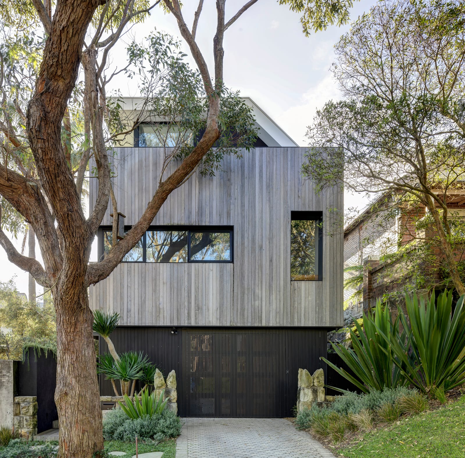 Quarry Box by MCK Architects. Photography by Brett Boardman. Street facade of timber clad, double-storey home. Aged timber clad on second story and black clad on ground floor. Stone dividing walls and green plants.