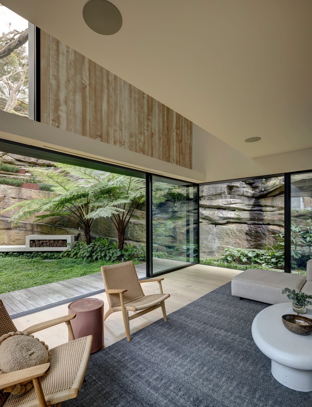 Quarry Box by MCK Architects. Photography by Brett Boardman. Living space with grey rug, two wicker armchairs and white round coffee table. Pale timber floors. Overlooking stone wall and green garden. 