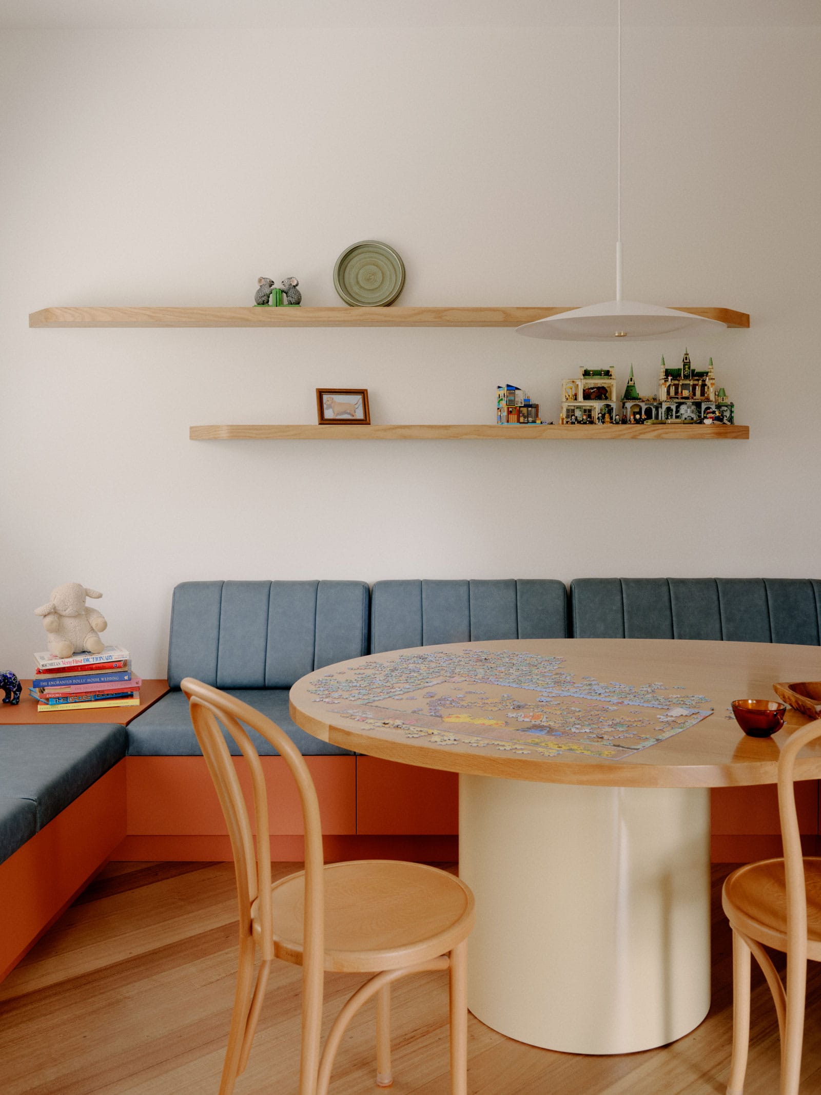 Selby House by Placement. Photography by Tom Ross. Integrated bench seating with clay coloured base and blue upholstery. Timber floors, dining table and dining chairs. Floating timber shelves above benches on white walls. 