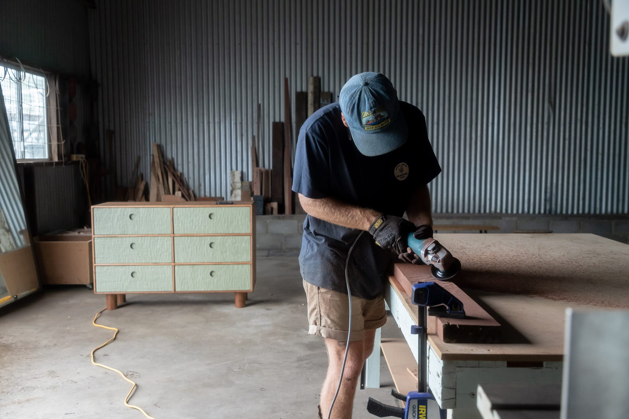 Two Blue Boys. Copyright of Two Blue Boys. Man uses electronic tool on piece of timber in a workshop. Timber and seafoam coloured sideboard in background. 