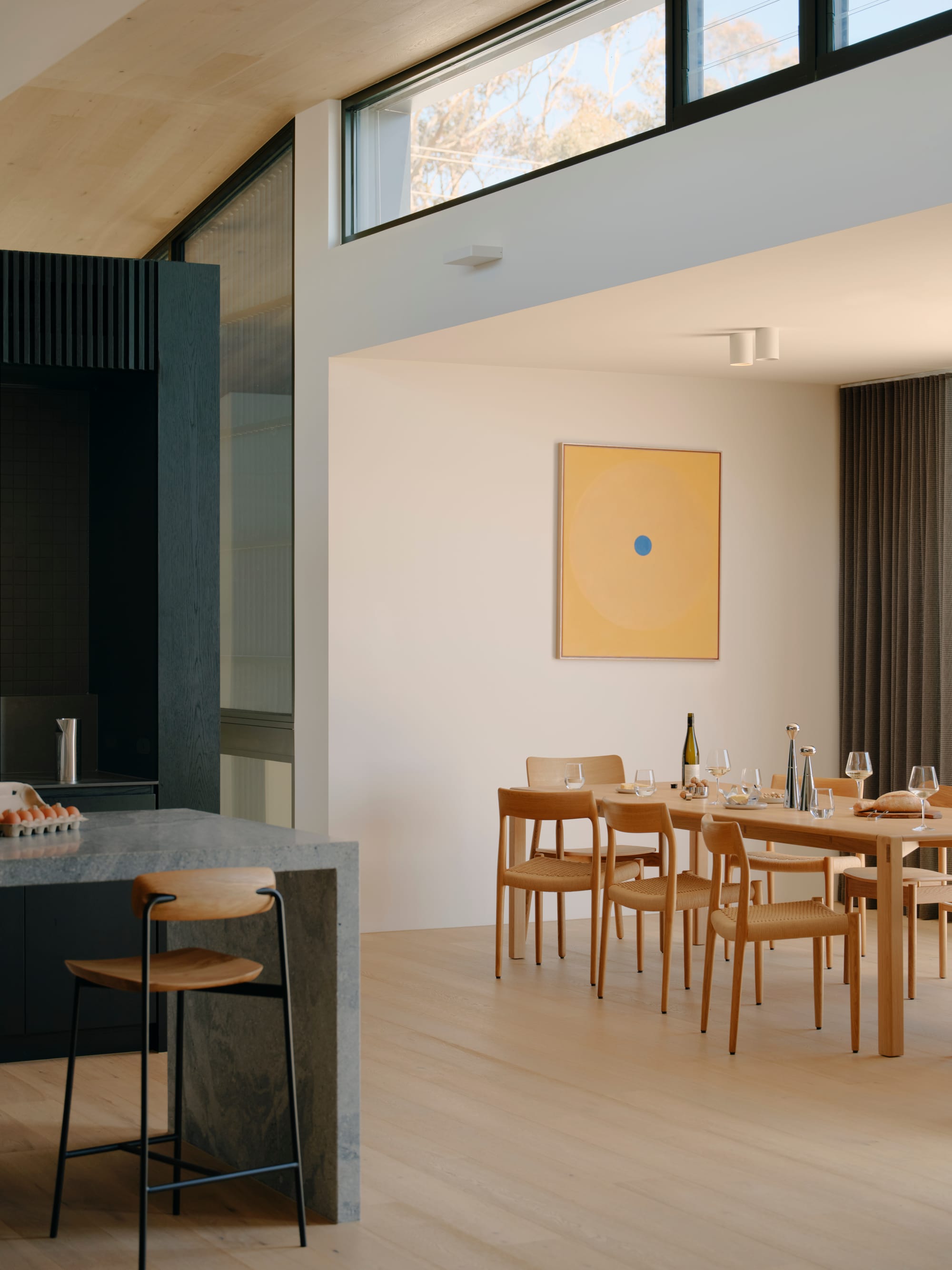 Courtside House by Tom Robertson Architects. Photography by Tom Ross. Kitchen and dining space with light timber floors, white walls and minimalistic timber chairs and stools. Granite waterfall benchtop to left of image. 